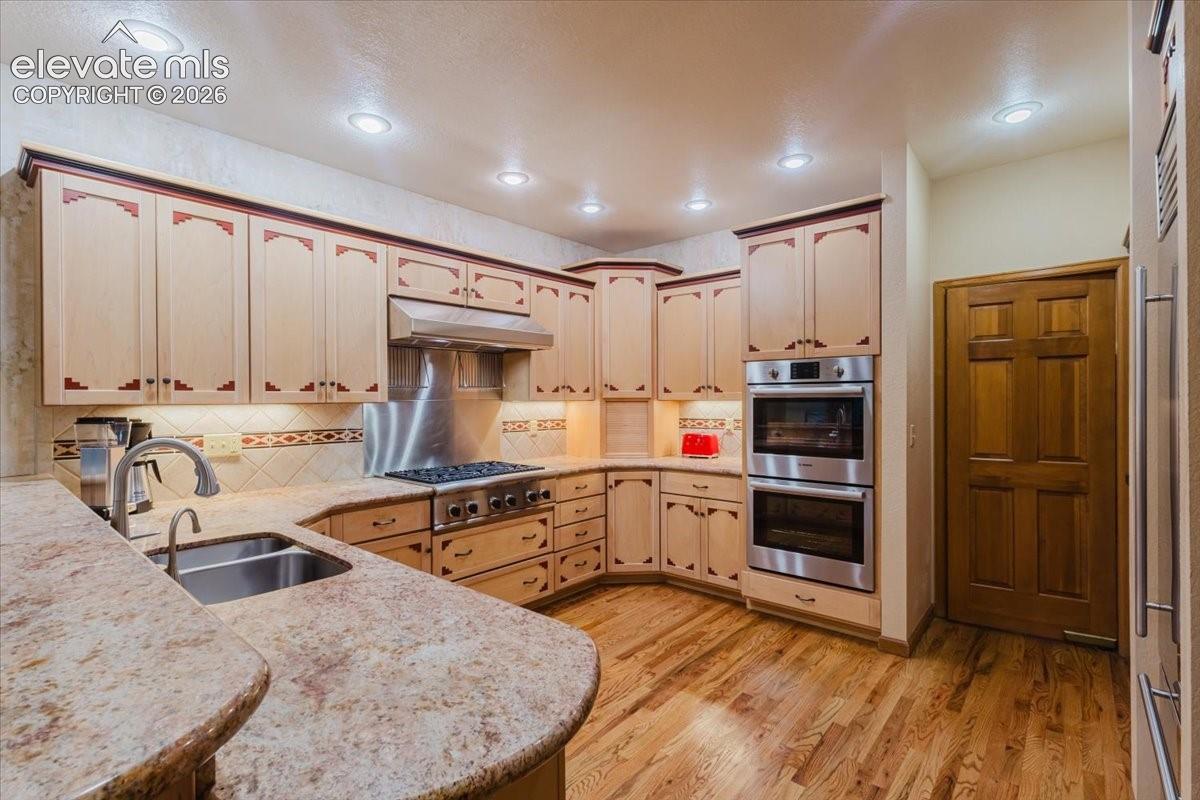 Kitchen featuring hard wood flooring, recessed lighting, double basin sink, stone countertops, and stainless steel appliances including a gas range, hood, and double wall ovens