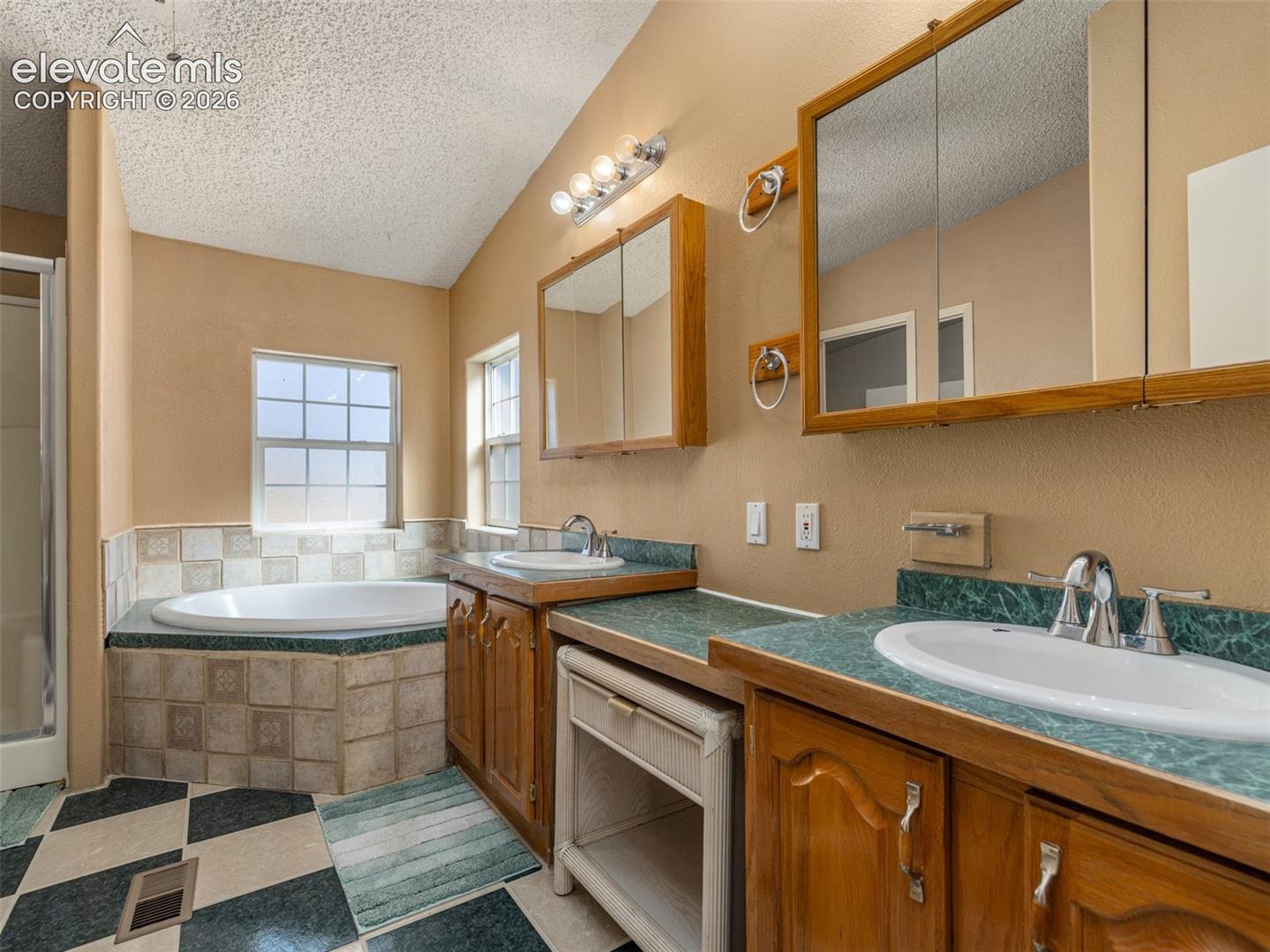 Dual-sink vanity with wood-finish cabinetry, solid surface countertops, two mirrored medicine cabinets, and a tiled built-in soaking tub
