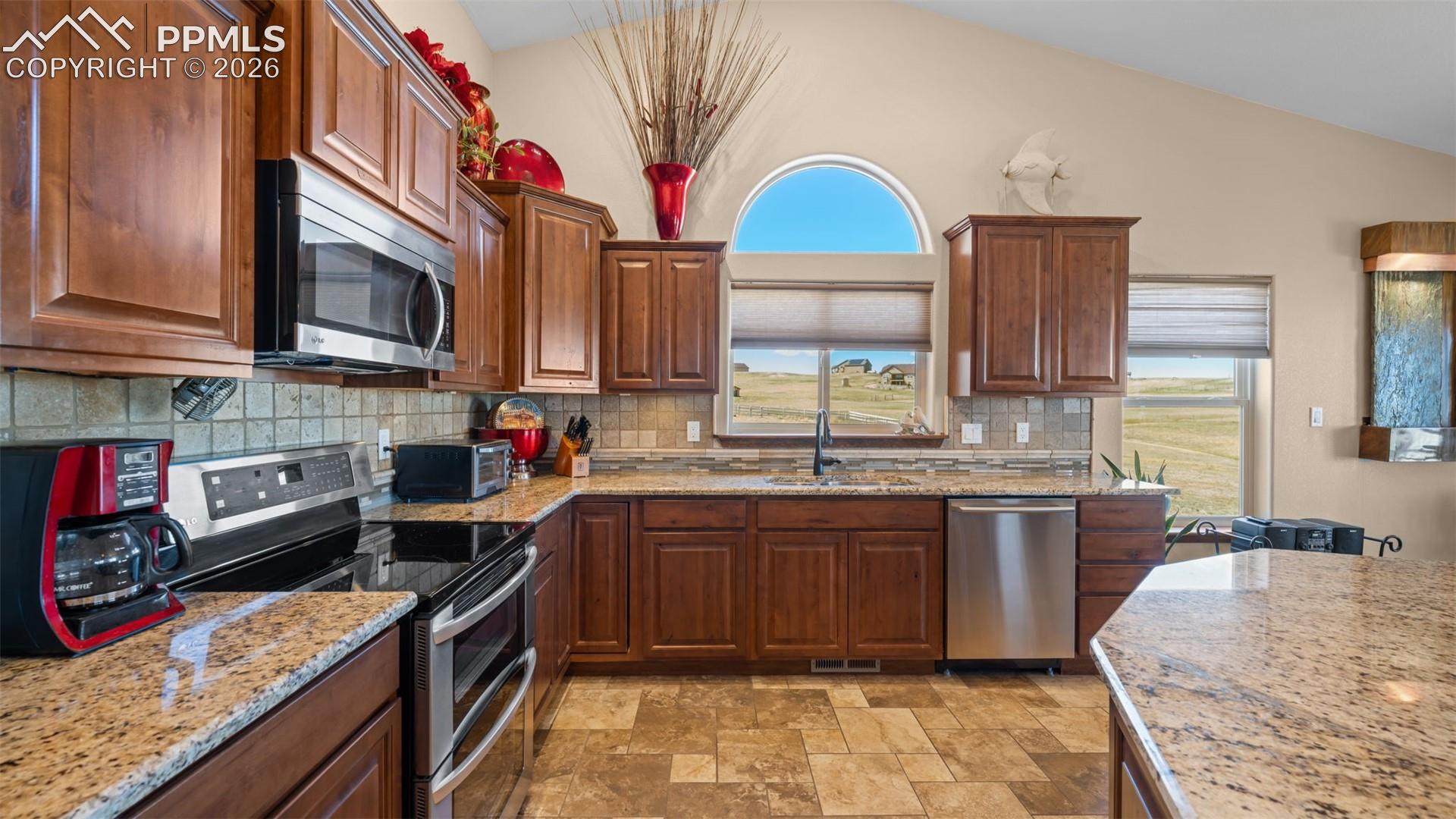 Kitchen with stainless steel appliances, stone finish flooring, lofted ceiling, light stone counters, and backsplash