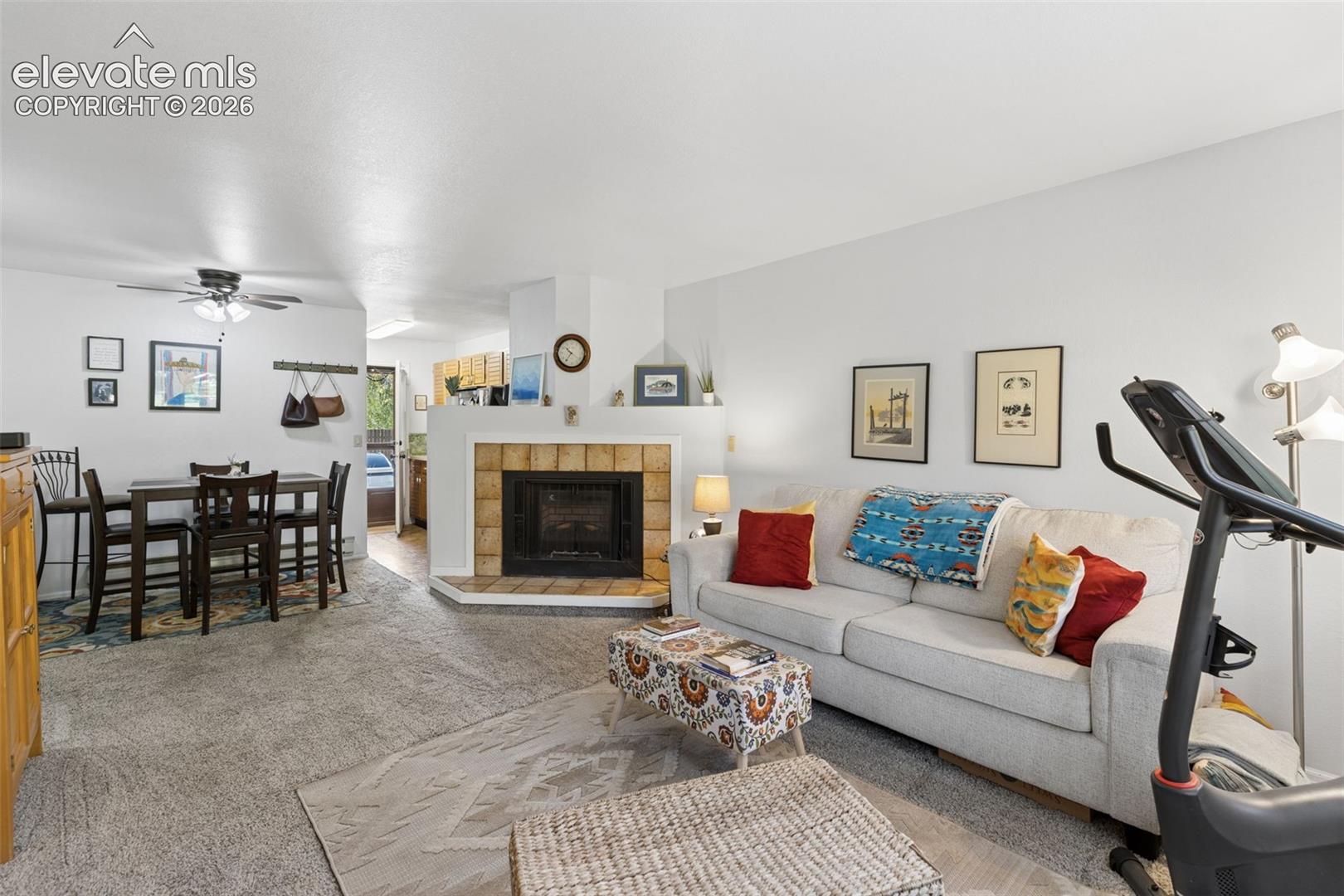Fireplace with a tiled surround, light-colored carpeting, and a ceiling fan with light fixture