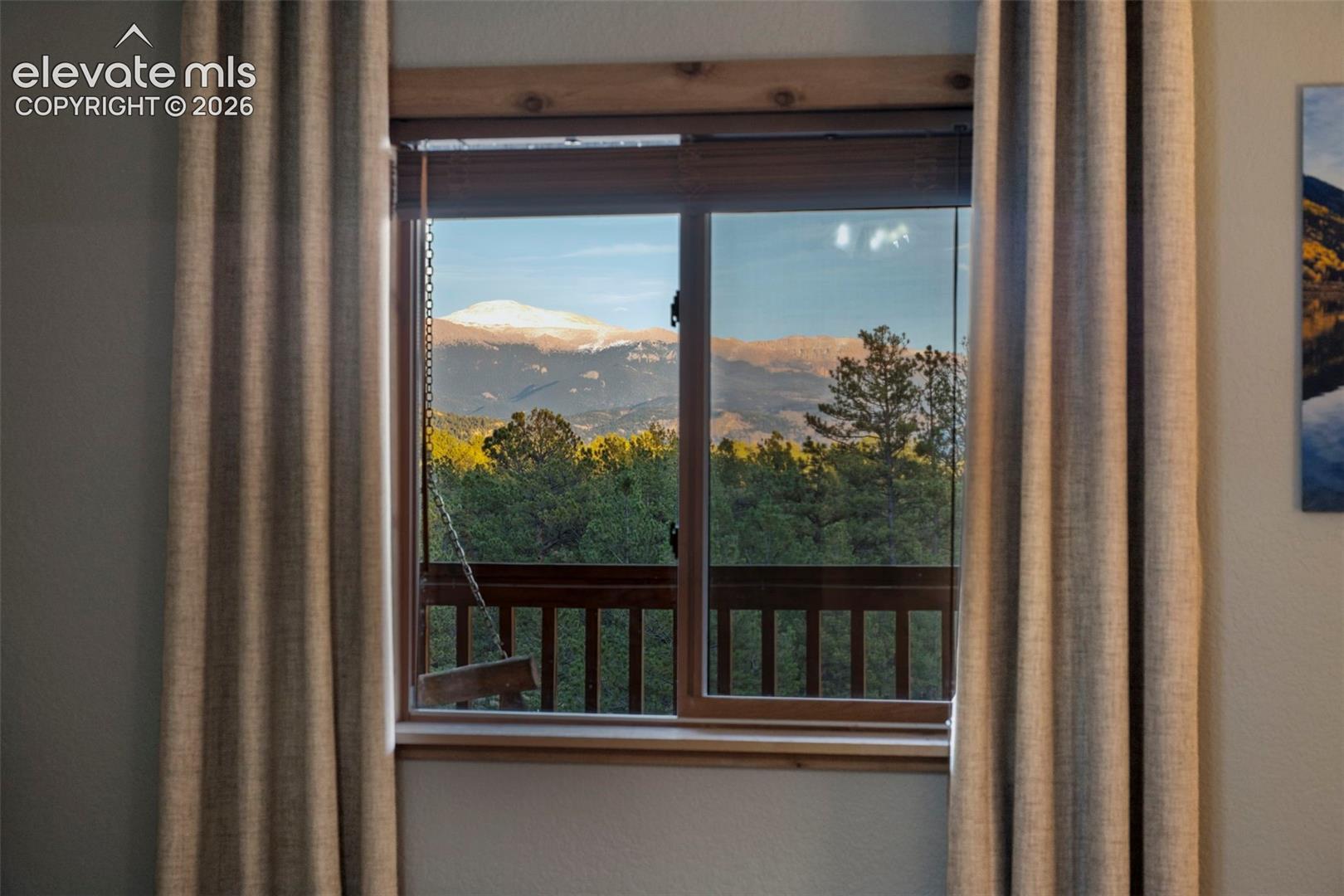Horizontal sliding window featuring a wood trim frame, revealing a landscape of mountain peaks, evergreen trees, and a wooden deck railing
