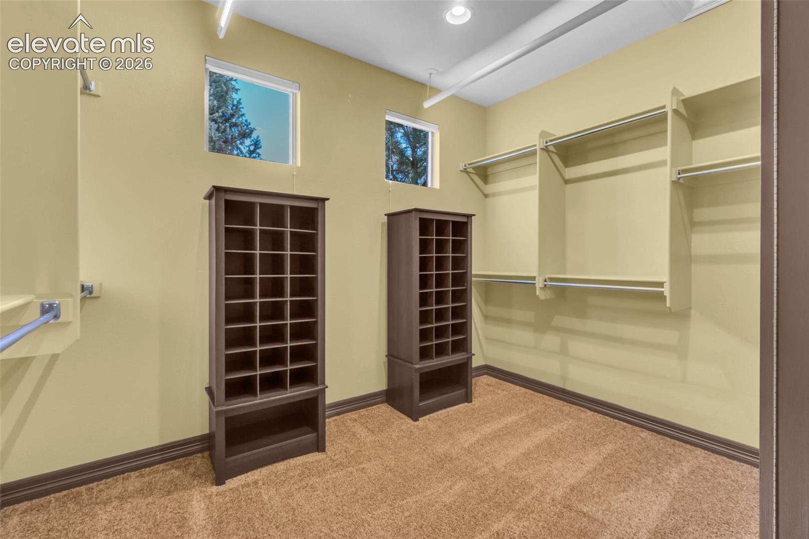 Walk-in storage space featuring built-in shelving towers, wall-mounted hanging rods, recessed ceiling lighting, and two square windows