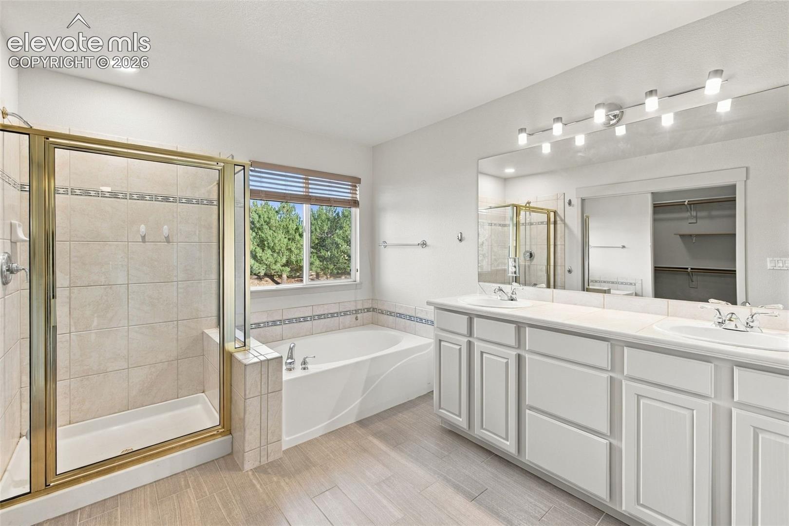 Dual-sink vanity with white cabinetry, soaking tub, glass-enclosed shower, wood-finish flooring, and wall-to-wall mirror