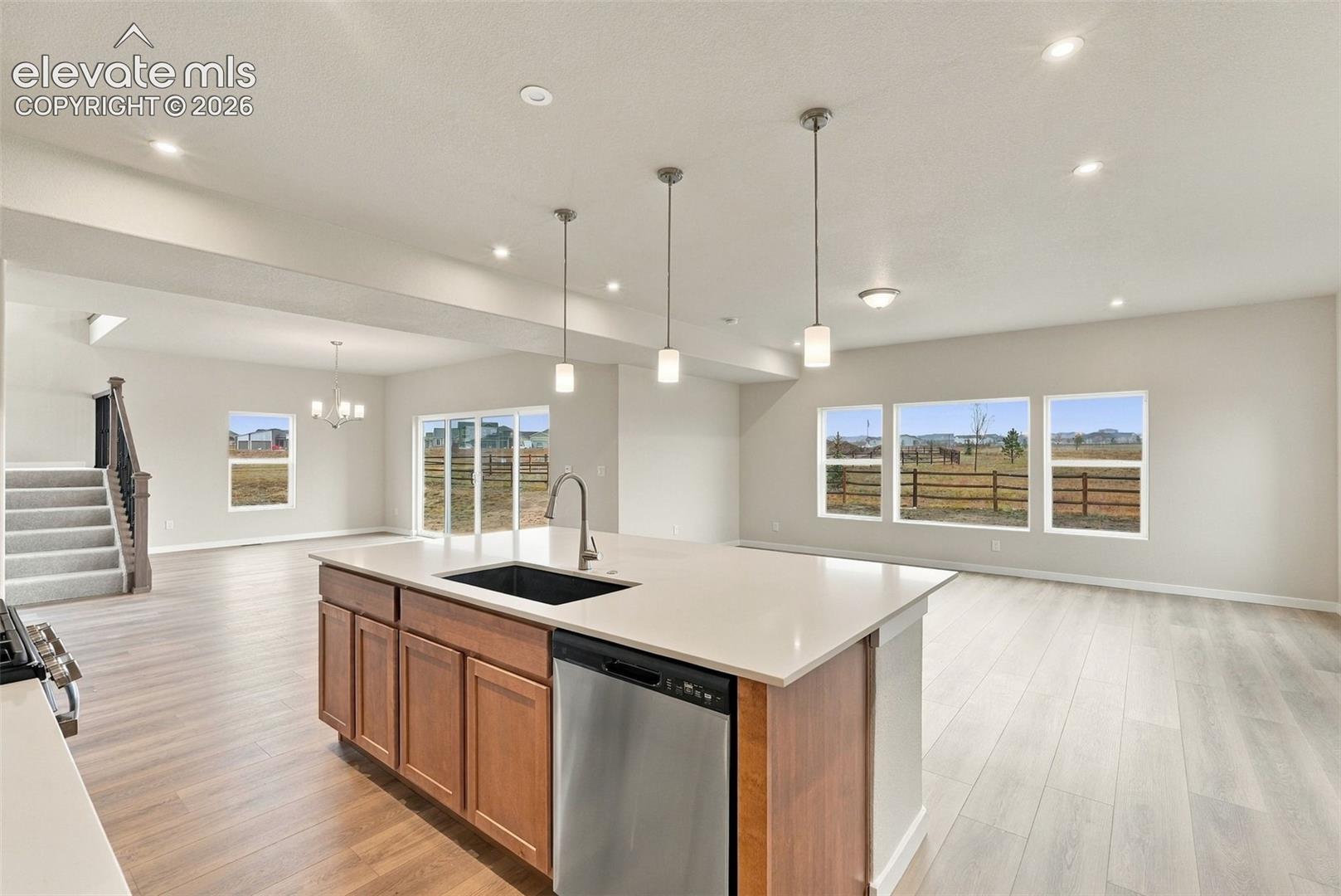 Kitchen island featuring a stone-surface countertop, an undermount sink with a gooseneck faucet, and stainless steel dishwasher, illuminated by three pendant lights, set against wood-finish flooring and multiple windows