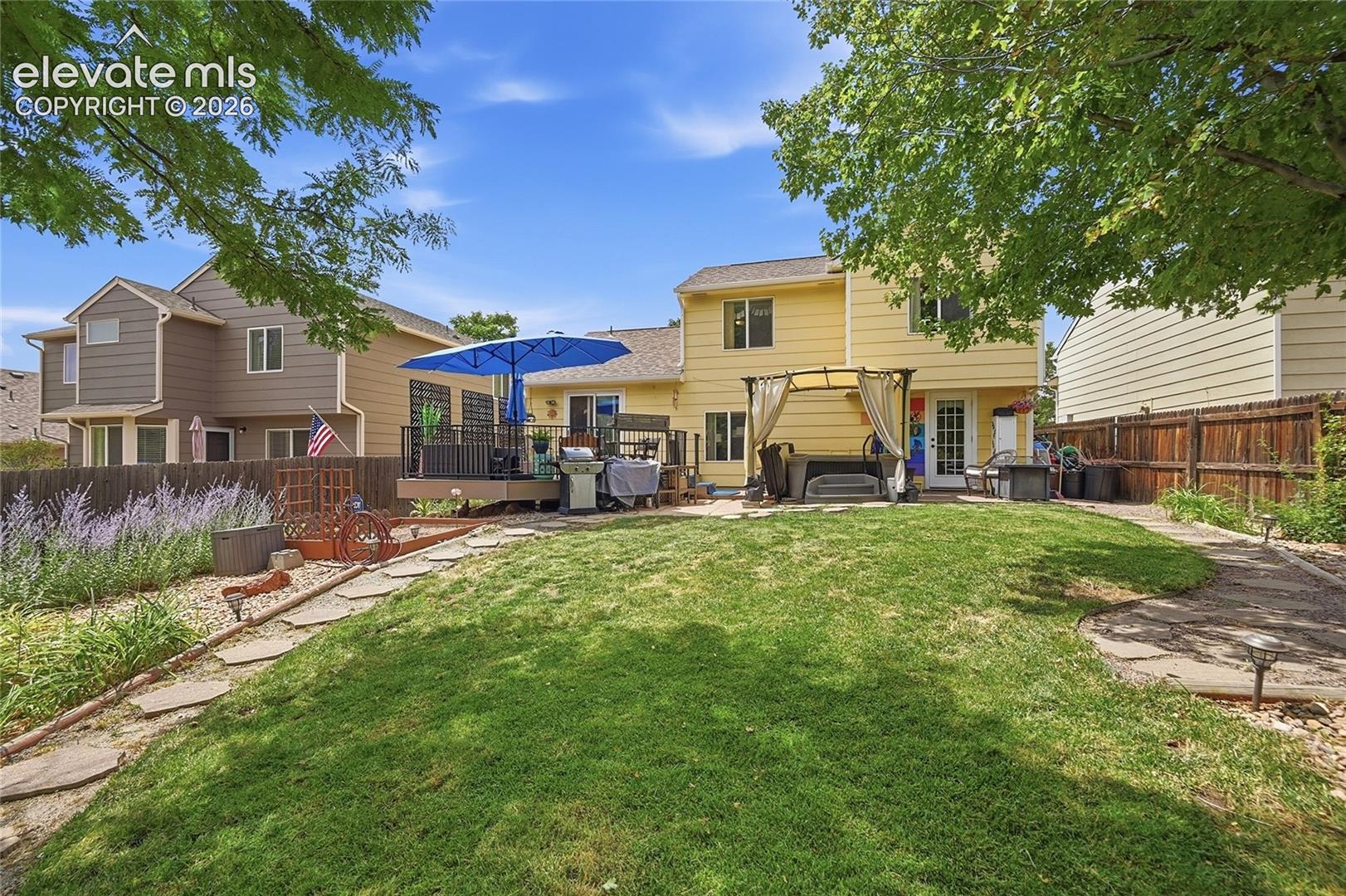Rear exterior featuring an elevated wood deck, stone-paver hardscaping, mature trees, and perimeter wood fencing