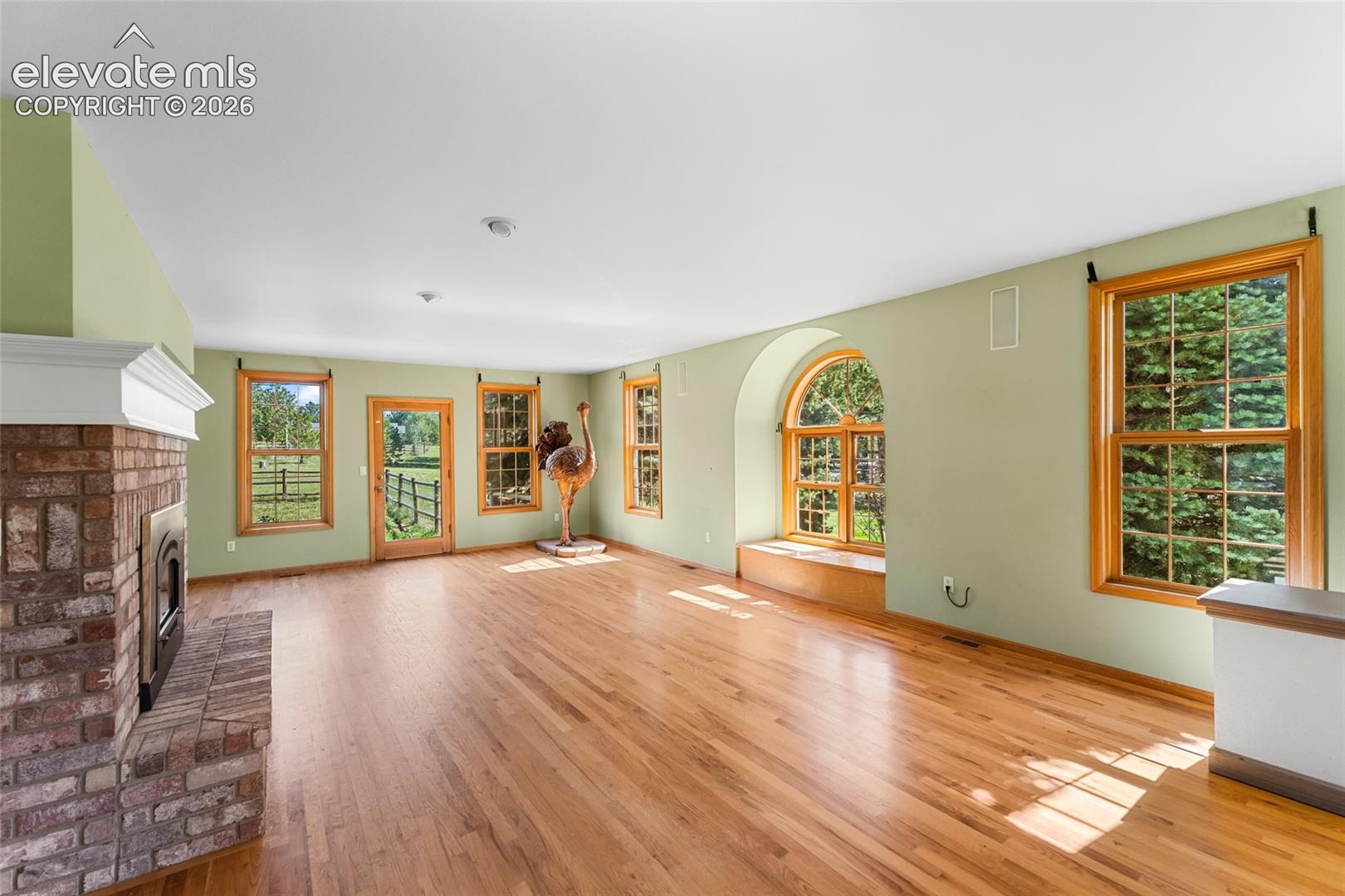 Brick fireplace with a white mantel, wood-finish flooring, multiple windows with wood trim, and an arched window with a built-in window seat