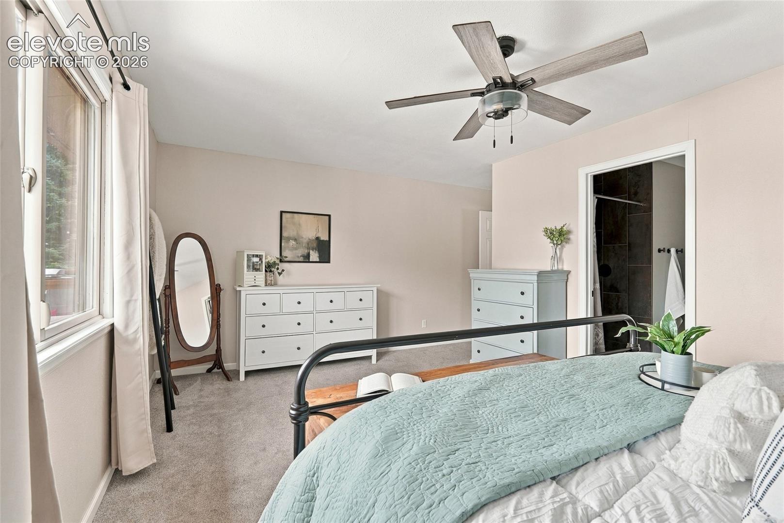Vaulted ceiling, ceiling fan with light fixture, window with trim, carpeted flooring, and an en-suite bathroom entry