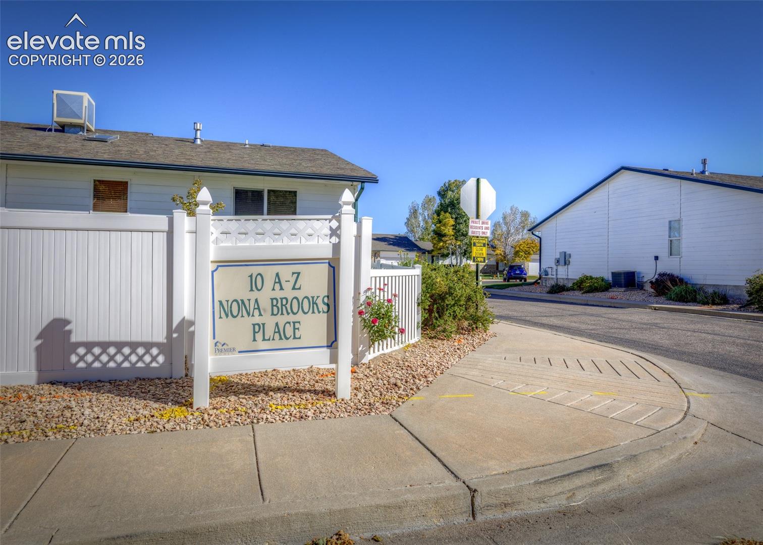 Freestanding property signage featuring decorative lattice trim, white vinyl fencing, concrete curbing, gravel landscaping, and asphalt paving