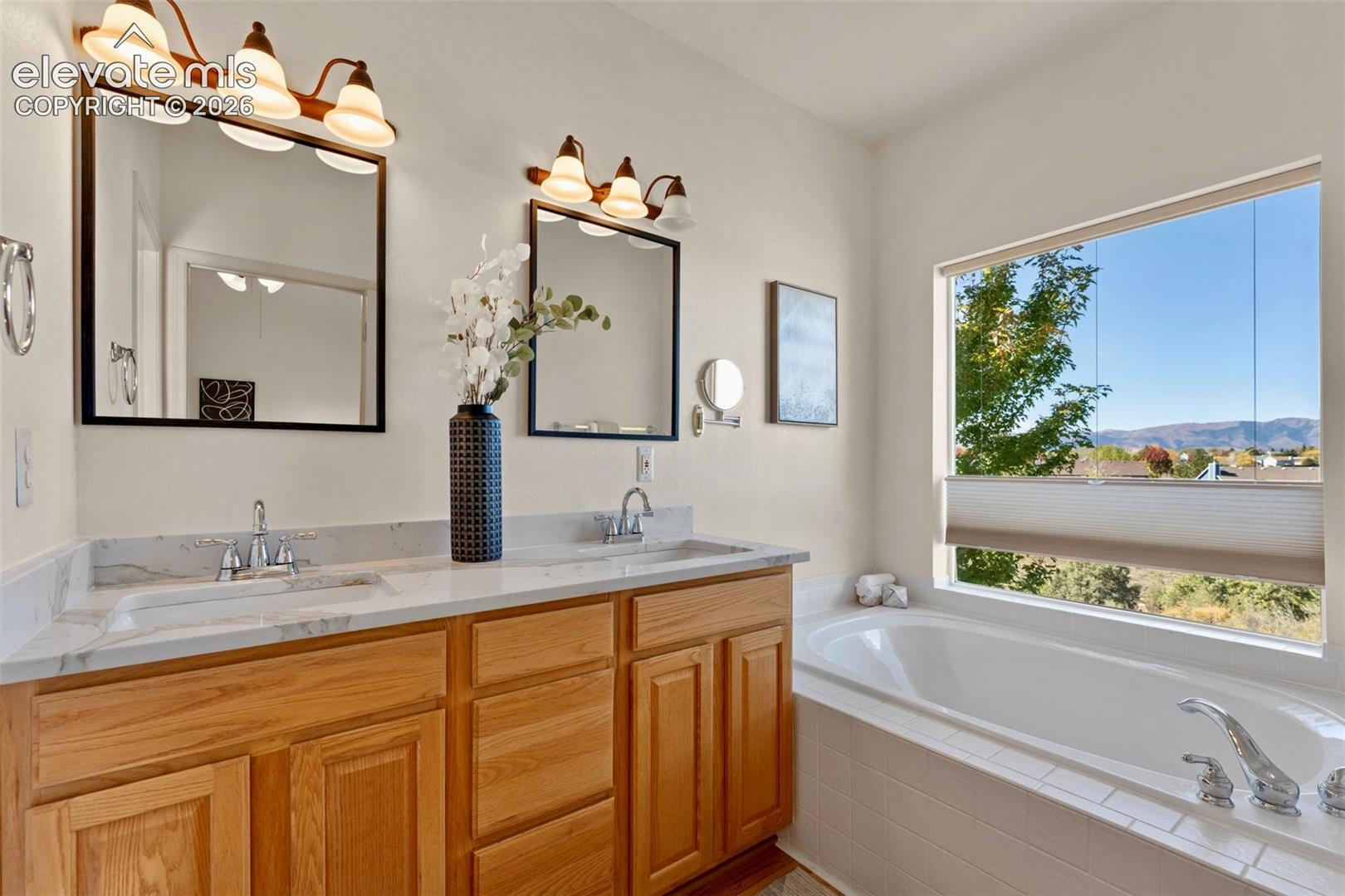 Double vanity with stone countertop, dual undermount sinks, wood-finish cabinetry, soaking tub with tile surround, and wall-mounted vanity lighting
