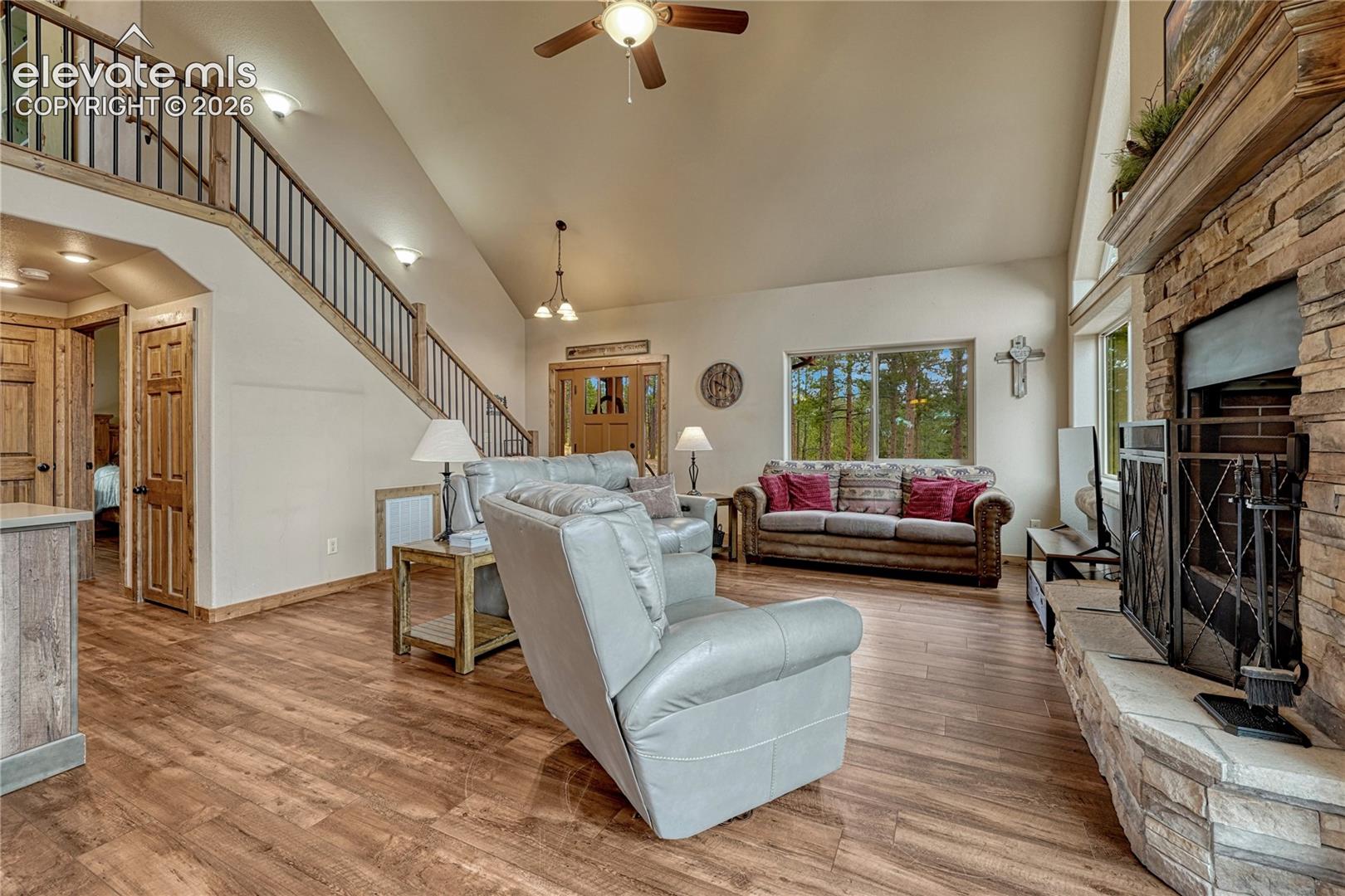 Vaulted ceiling featuring a ceiling fan, stone-surround fireplace, wood-finish flooring, and an open staircase with a wood and metal railing