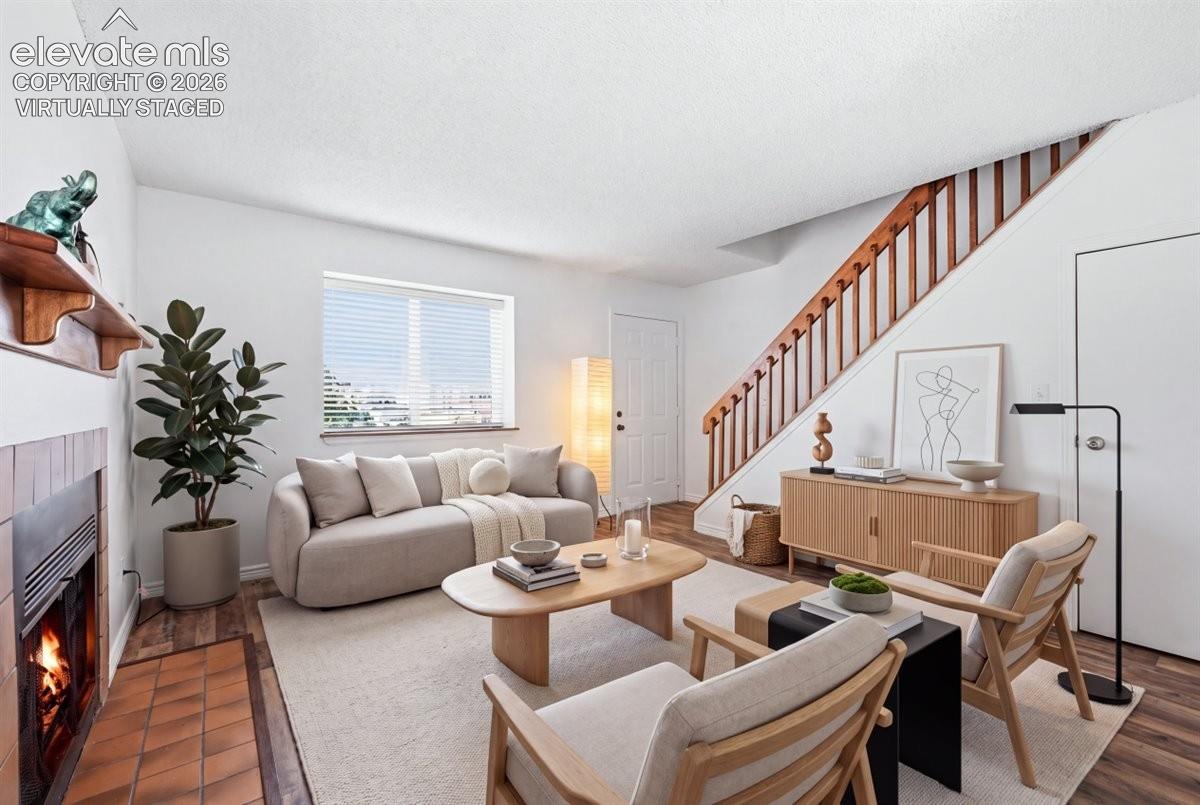 VIRTUALLY STAGED
Living area featuring wood-finish flooring, a fireplace with tile surround, a window with horizontal blinds, and a staircase with wood railing