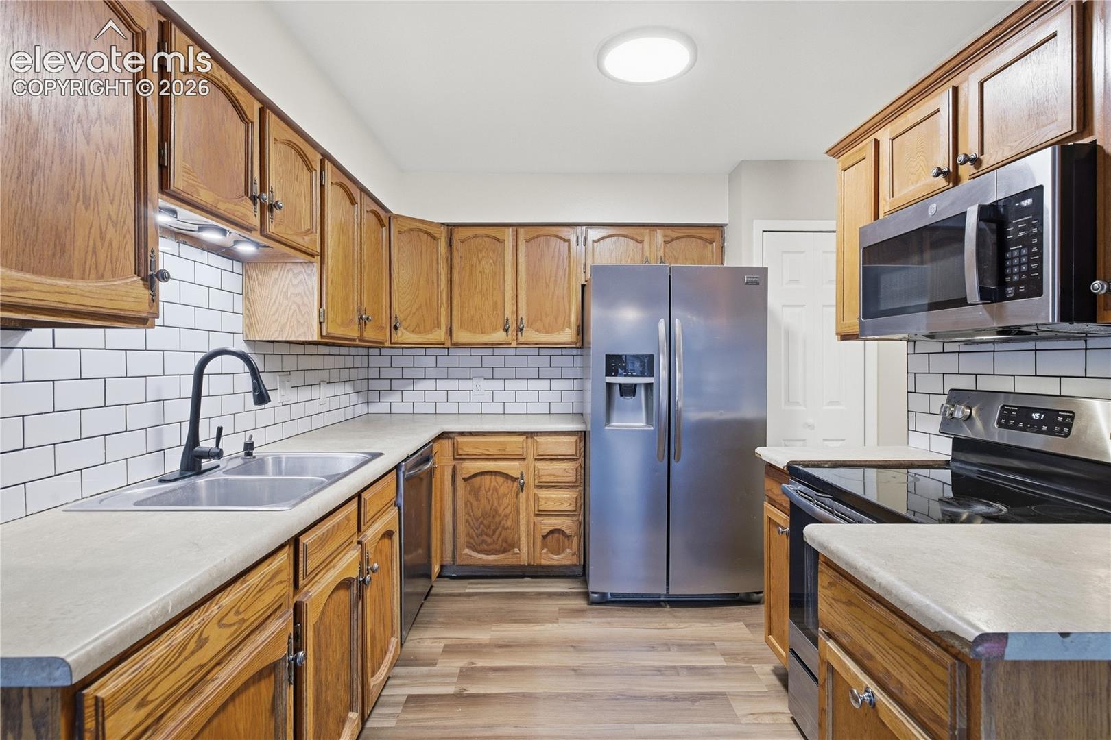 Kitchen featuring wood cabinetry, subway tile backsplash, stainless steel appliances, dual basin sink with gooseneck faucet, and wood-finish flooring