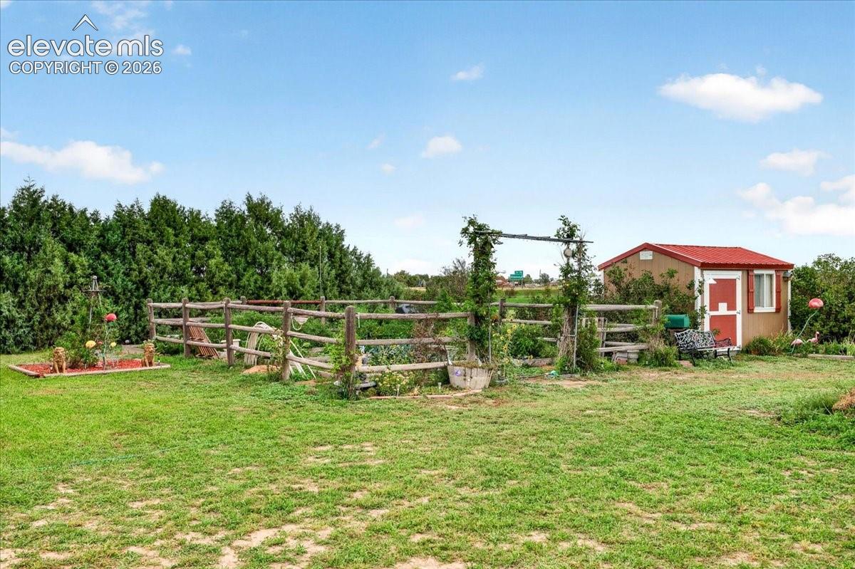 Expansive backyard featuring a rustic rail fence, a dedicated gardening area with a pergola, mature perimeter evergreen trees, and a detached storage structure with a metal roof