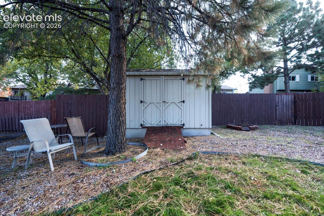 Exterior storage structure with double-door entry, concrete foundation, and ramp, set within a fenced yard featuring mature trees