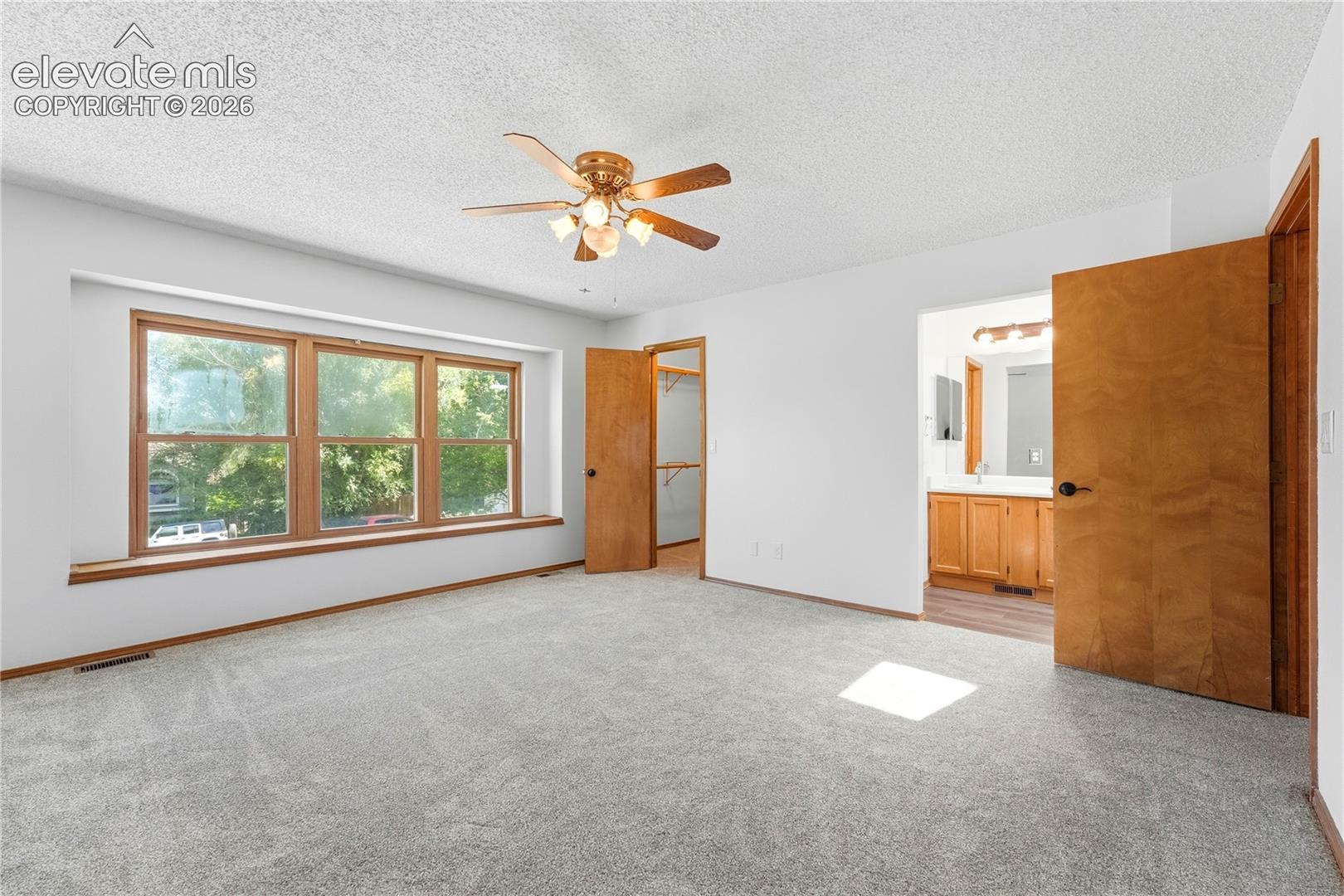 Spacious room featuring light gray carpeting, a wide window with faux wood vinyl trim, and a ceiling fan with lighting