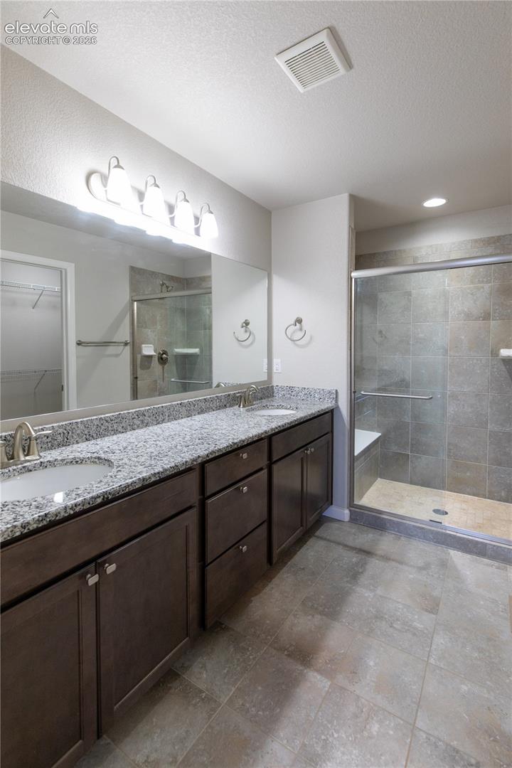 Dual-sink vanity featuring dark-finish cabinetry, stone-patterned countertop, full-width mirror, and a walk-in shower with tiled surround and built-in bench
