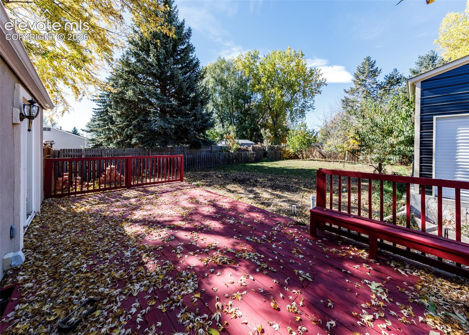 Elevated wood deck featuring red railing and integrated bench seating, overlooking a fenced backyard with mature trees