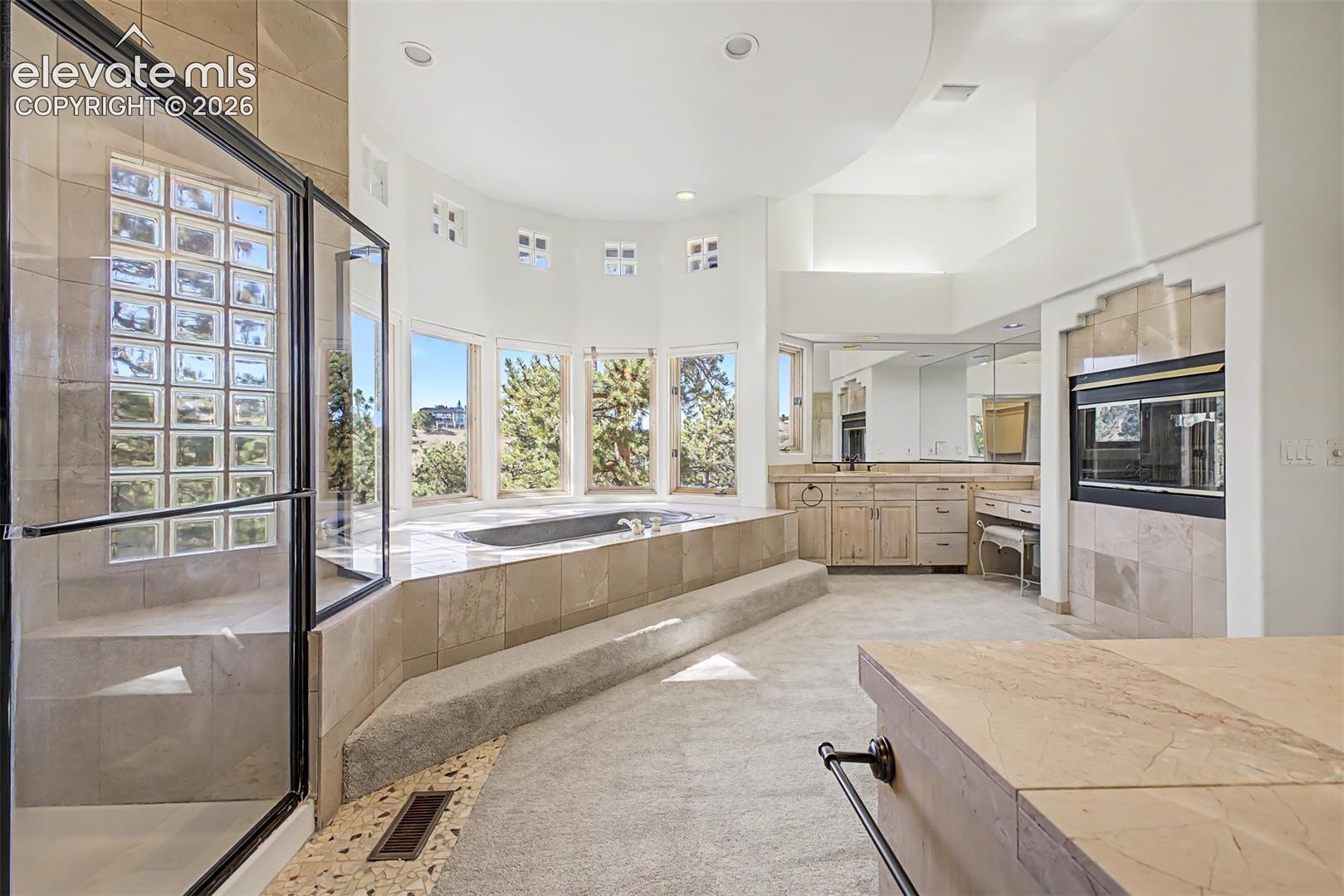Built-in soaking tub, glass-enclosed shower, dual vanity with undermount sinks, wood-finish cabinetry, and integrated wall oven