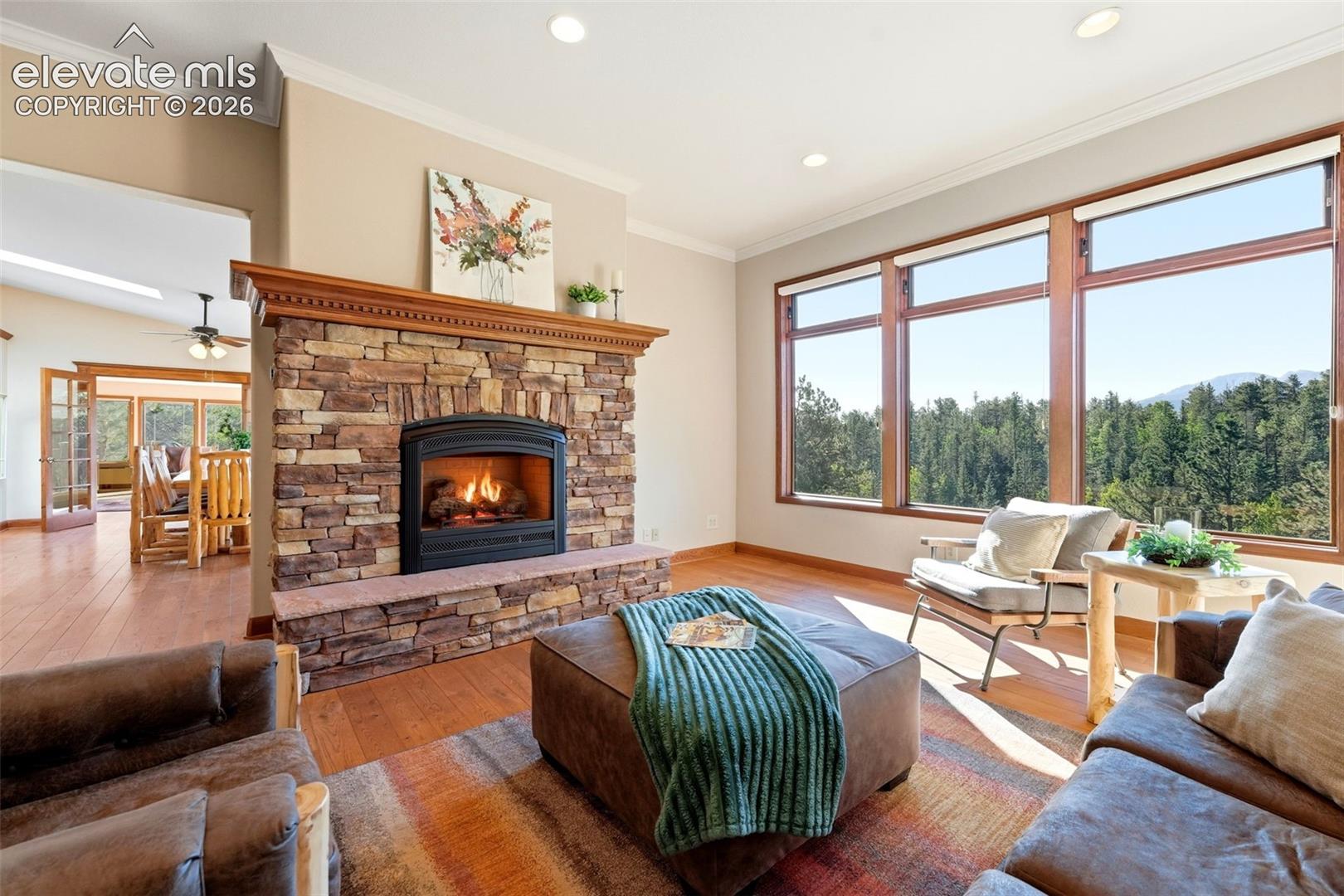Floor-to-ceiling stone fireplace with a wood mantle, wood-finish flooring, recessed lighting, and large windows with wood frames
