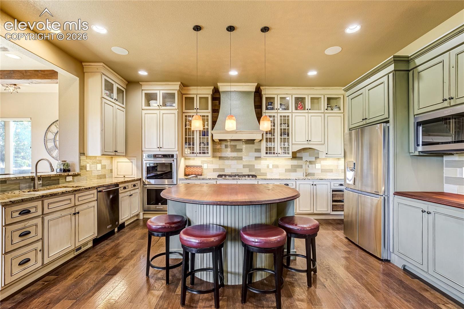 Soft-close Schroll cabinetry in cream with cocoa glaze + sage rises to the ceiling, accented by crown molding, glass displays + under-cabinet lighting. Granite counters complement a butcher-block island.