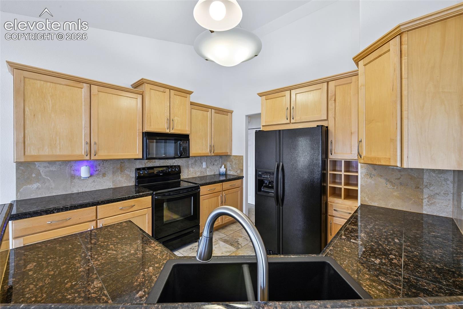 Kitchen featuring light wood cabinetry, dark speckled countertops, stone-finish backsplash, and a black double-basin sink with a gooseneck faucet