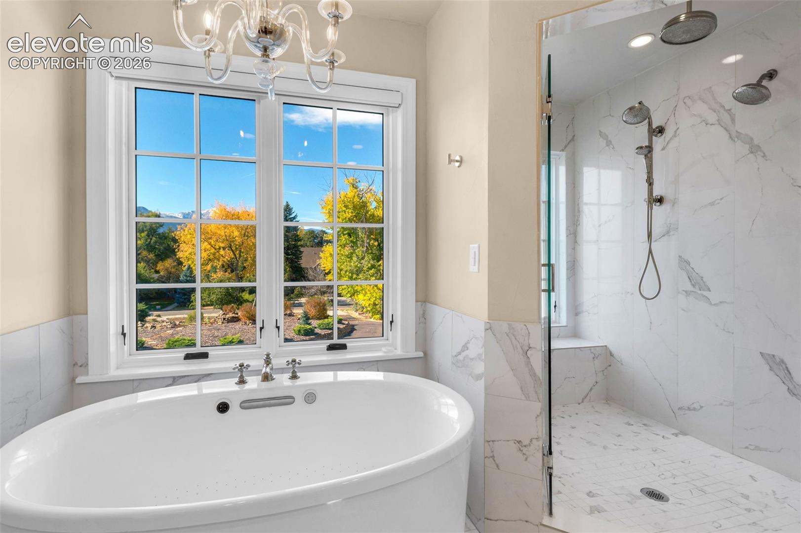 Oval freestanding soaking tub with chrome fixtures, glass-enclosed walk-in shower featuring tile surrounds, multi-head shower system, and crystal-style chandelier
