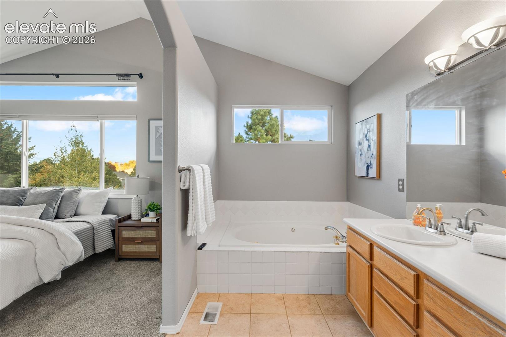 Vaulted ceilings, drop-in soaking tub with white tile surround, vanity with wood-finish cabinetry, and large windows with natural light