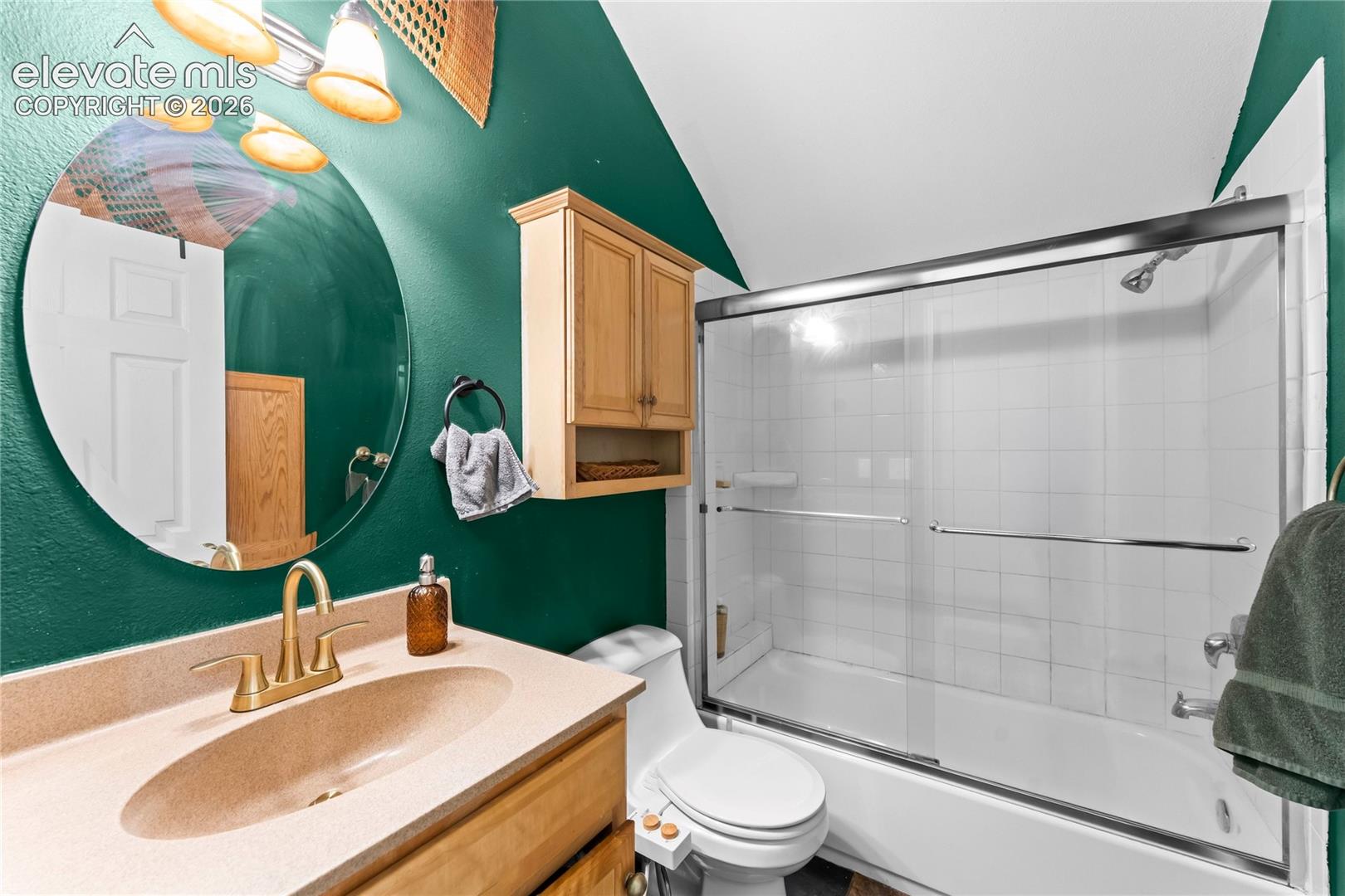 Full bathroom featuring a vanity with integrated sink, brass-finish faucet, wall-mounted cabinet, circular mirror, and a tub-shower combination with sliding glass doors