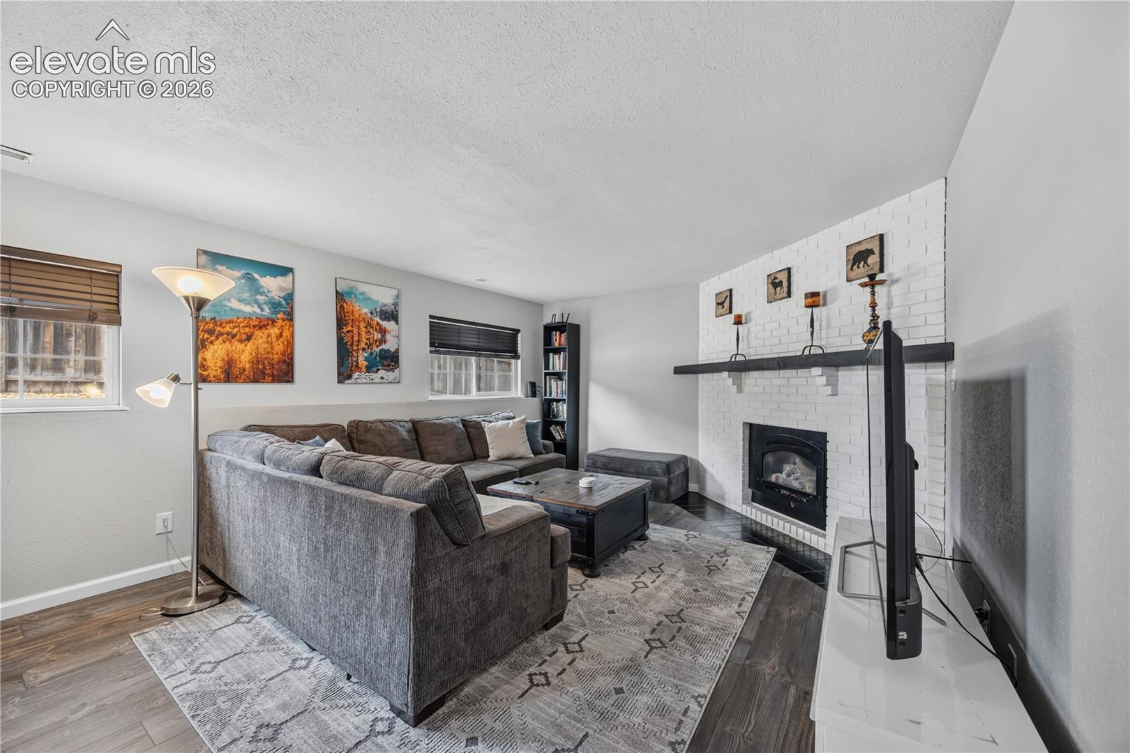 Living area featuring a floor-to-ceiling painted brick fireplace, contrasting dark mantel, wood-finish flooring, and natural light from multiple windows
