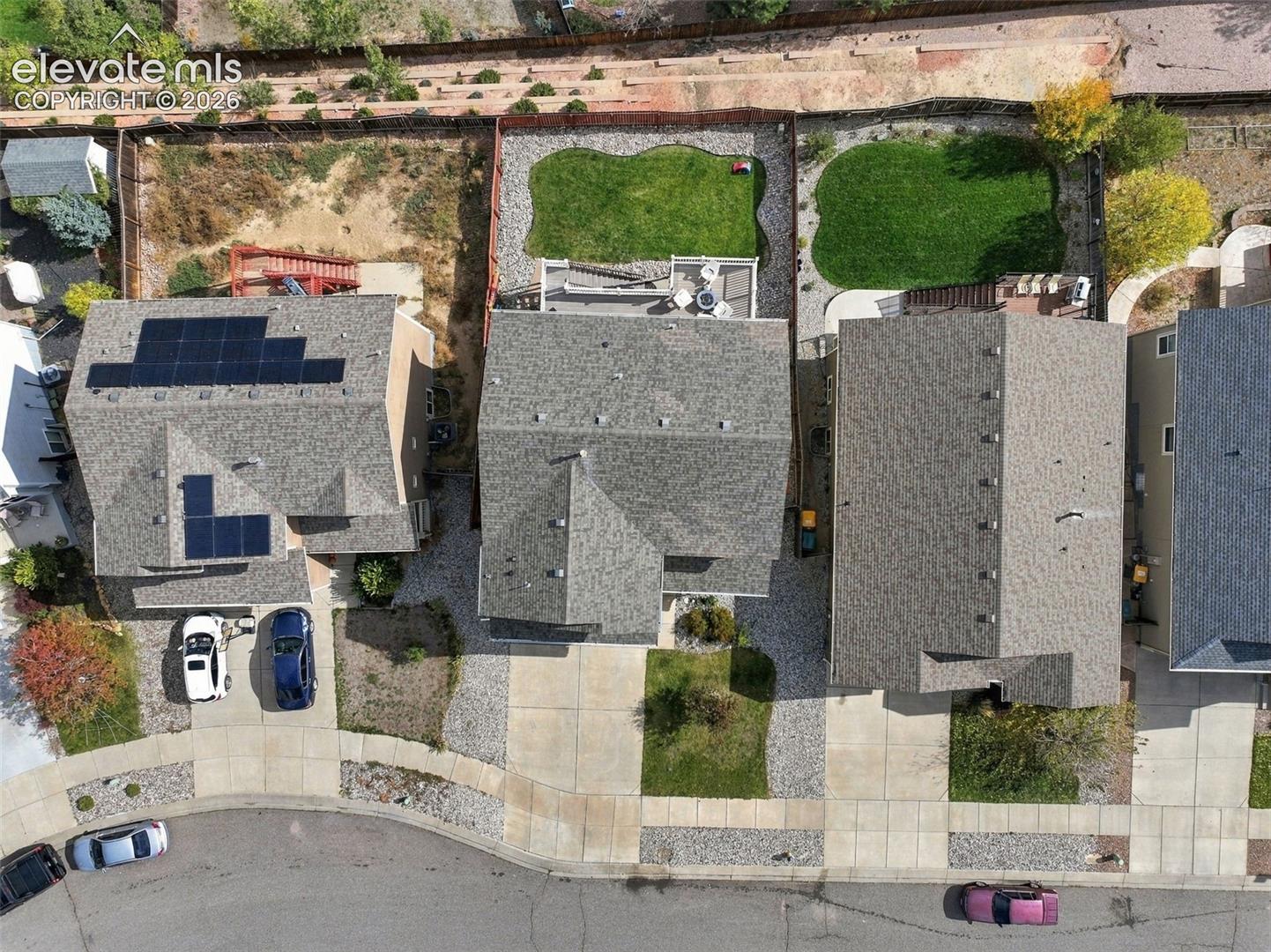 Aerial perspective of property featuring a composition shingle roof with integrated solar panels, paved driveway, and a rear fenced yard with a manicured lawn and multi-level deck