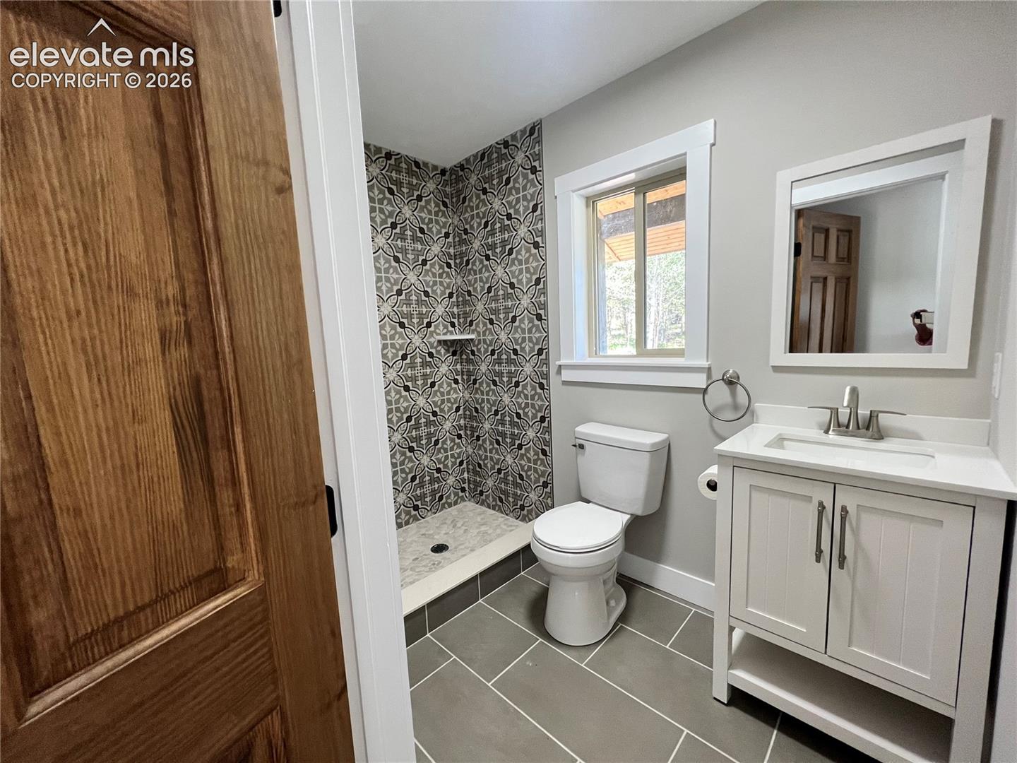 Walk-in shower with decorative tiled surround, single-sink vanity with white cabinetry, square mirror, and wood-finish door