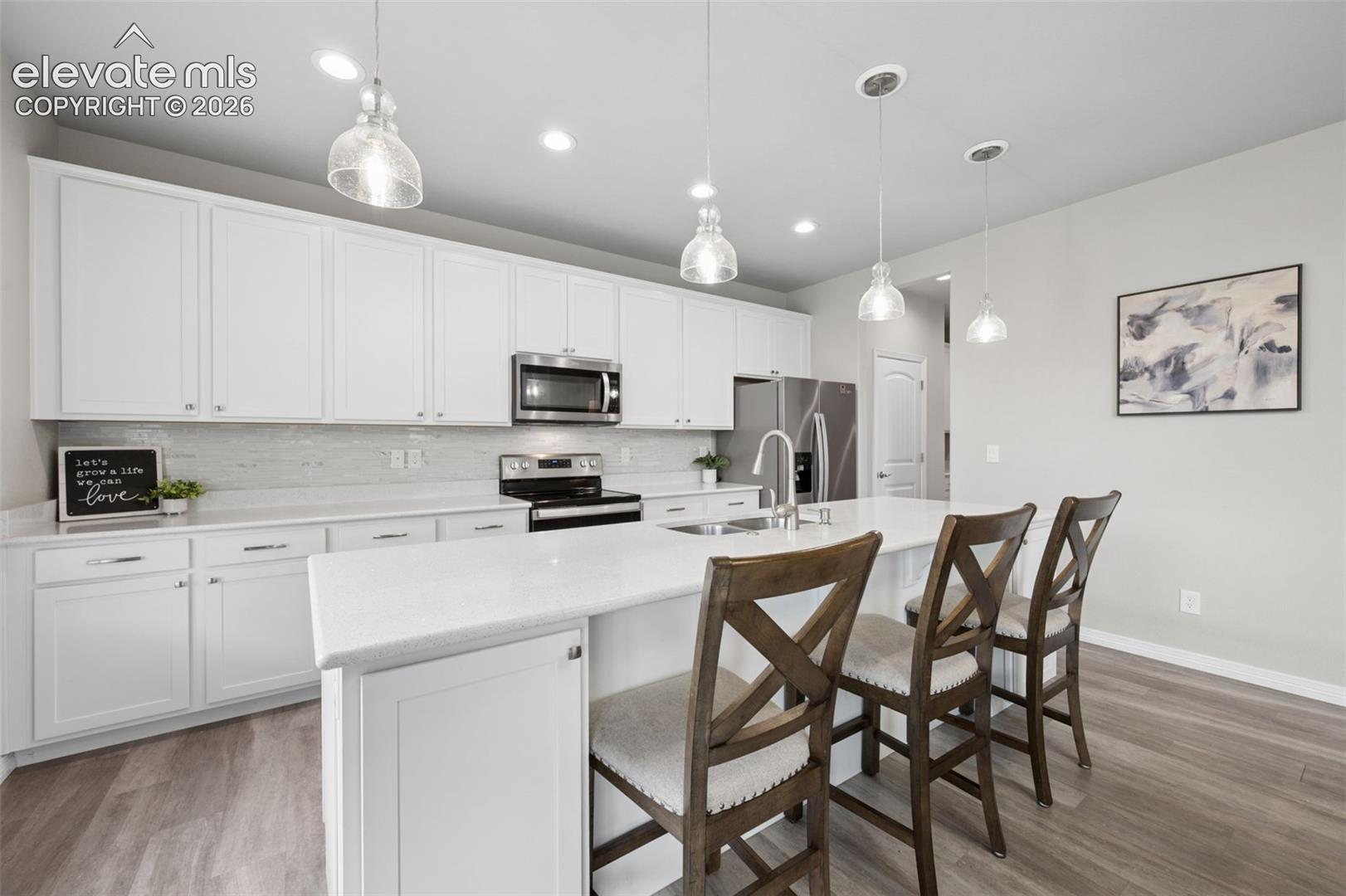 White cabinetry, light-toned backsplash, stainless steel appliances, large kitchen island with counter seating, and wood-finish flooring