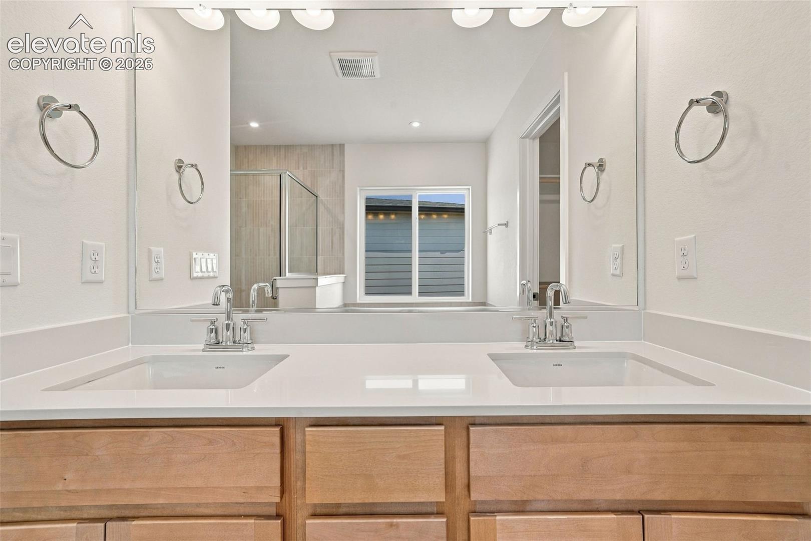 Dual undermount sinks with chrome faucets, light wood-finish cabinetry, broad vanity mirror, and overhead vanity lighting