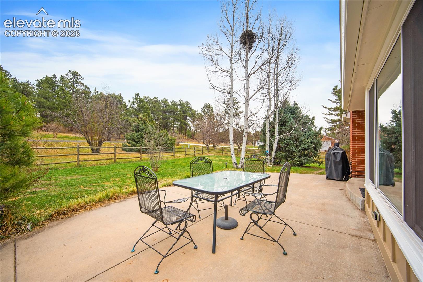 Concrete patio featuring a brick exterior facade, split-rail fencing, and mature trees