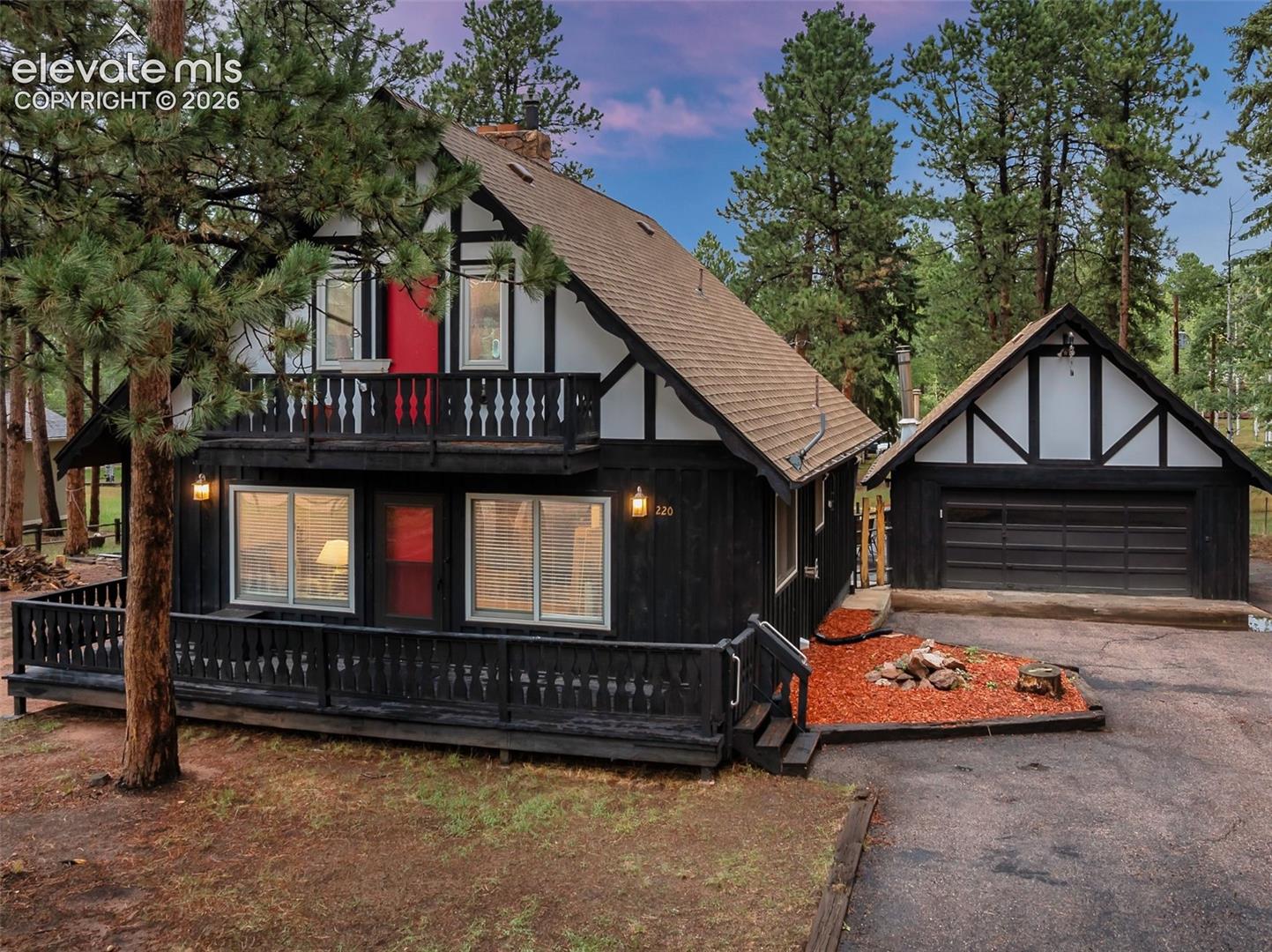Tudor-style exterior with black siding and white trim, featuring an upper-level balcony, a ground-level porch with wood railings, and an attached two-car garage
