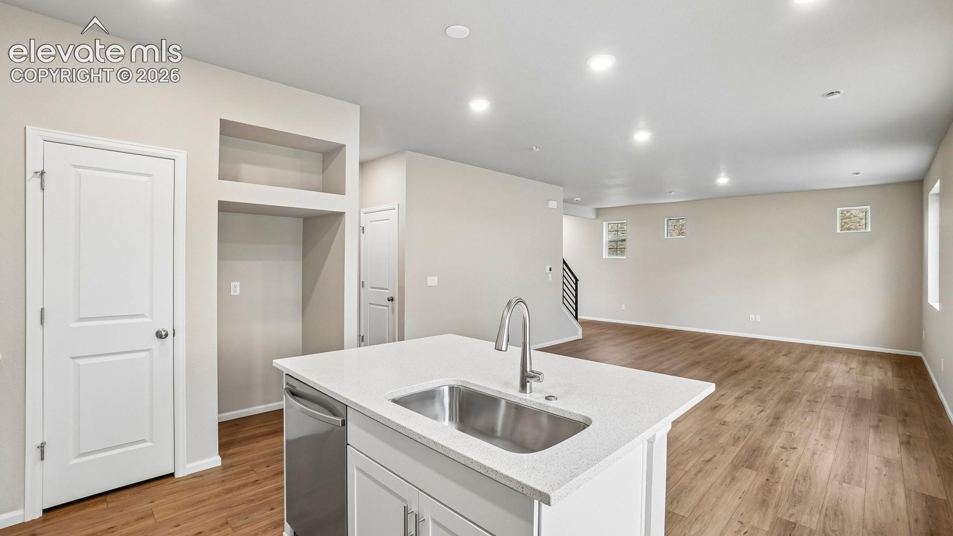 Open-concept kitchen and living area featuring a white cabinetry island with stainless steel undermount sink and chrome faucet, white quartz countertops, wood-finish flooring throughout, recessed ceiling lighting, and ample natural light from multiple windows
*photos of a model home*