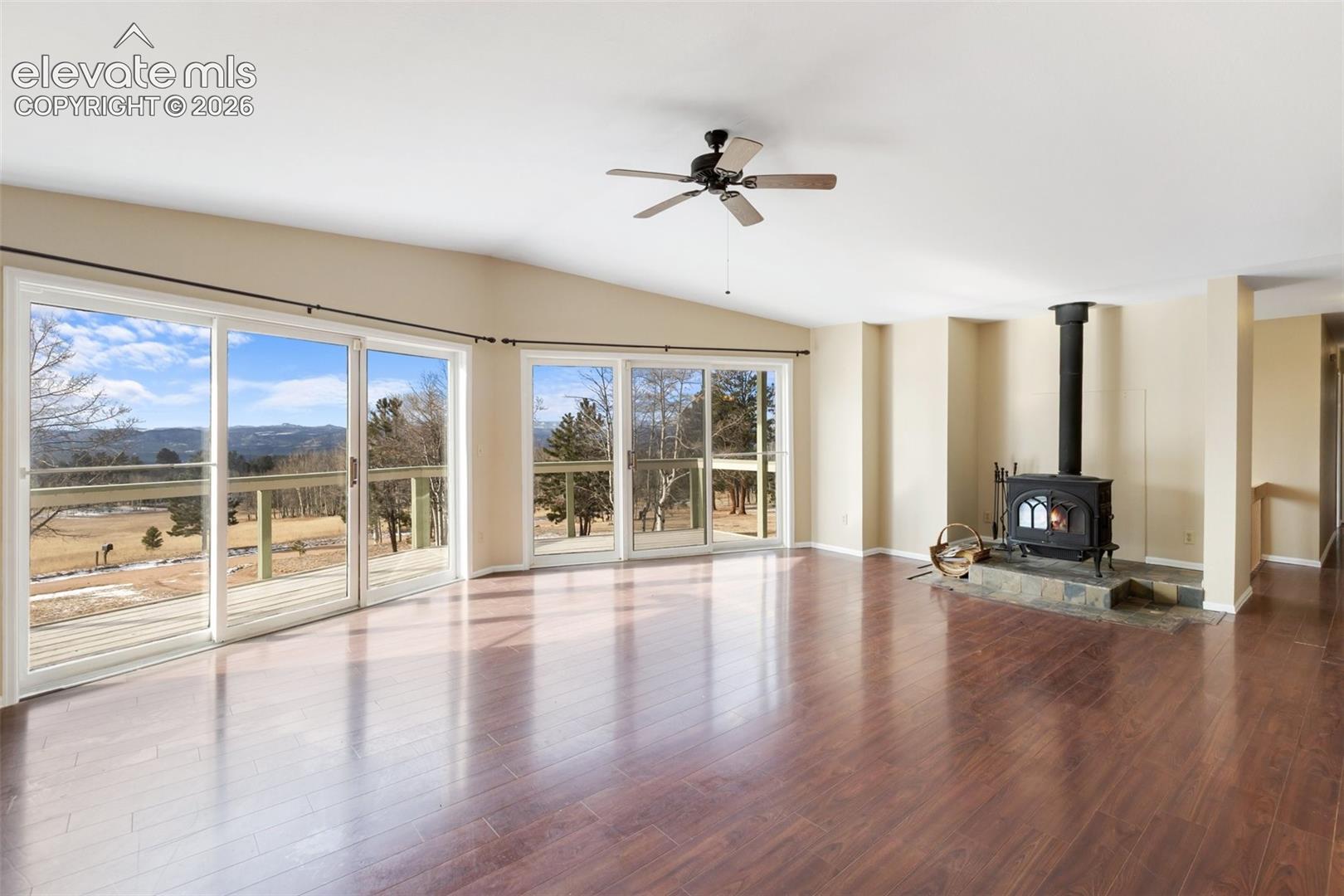 Vaulted ceiling with a ceiling fan, wood-finish flooring, wood-burning stove with a stone hearth, and multiple sliding glass doors leading to an exterior deck