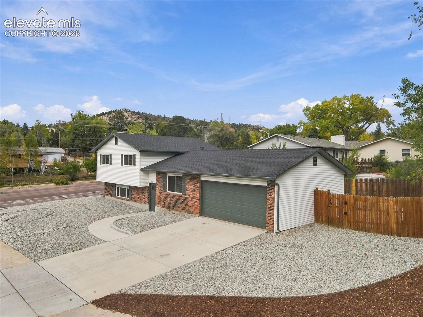 Split-level exterior featuring brick and siding facade, an attached two-car garage, gravel landscaping, a concrete driveway, and a wooden privacy fence