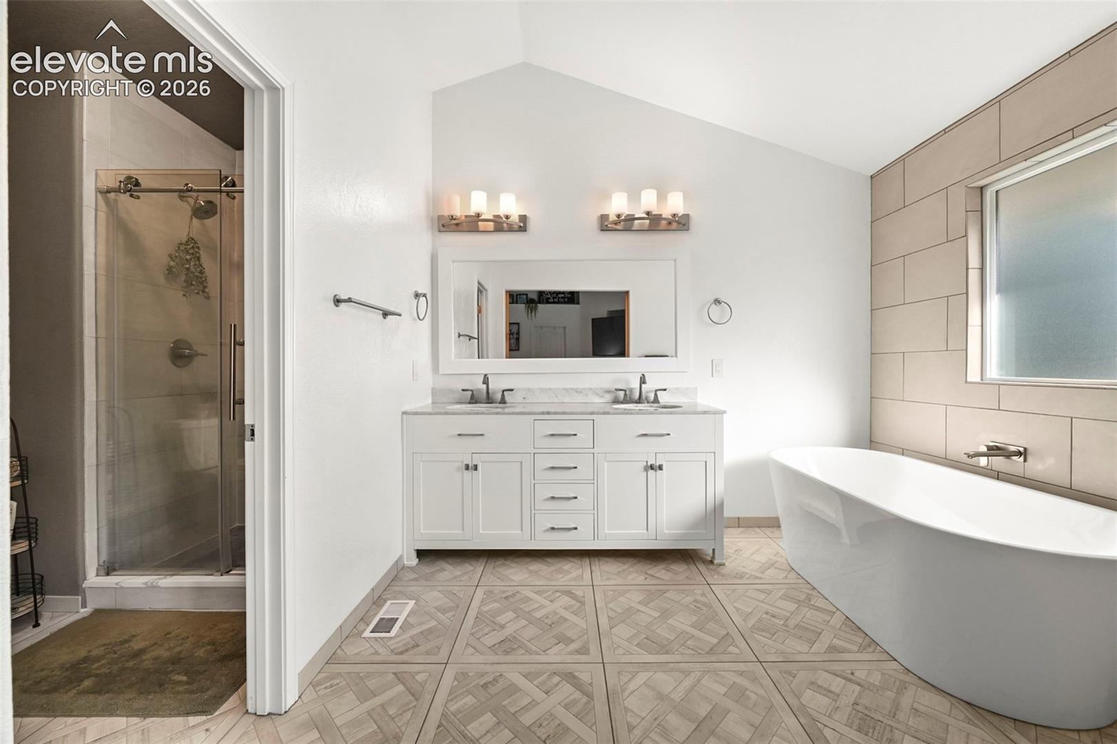 Double vanity with white cabinetry, freestanding soaking tub, tiled shower with glass enclosure, tiled accent wall, and patterned tile flooring