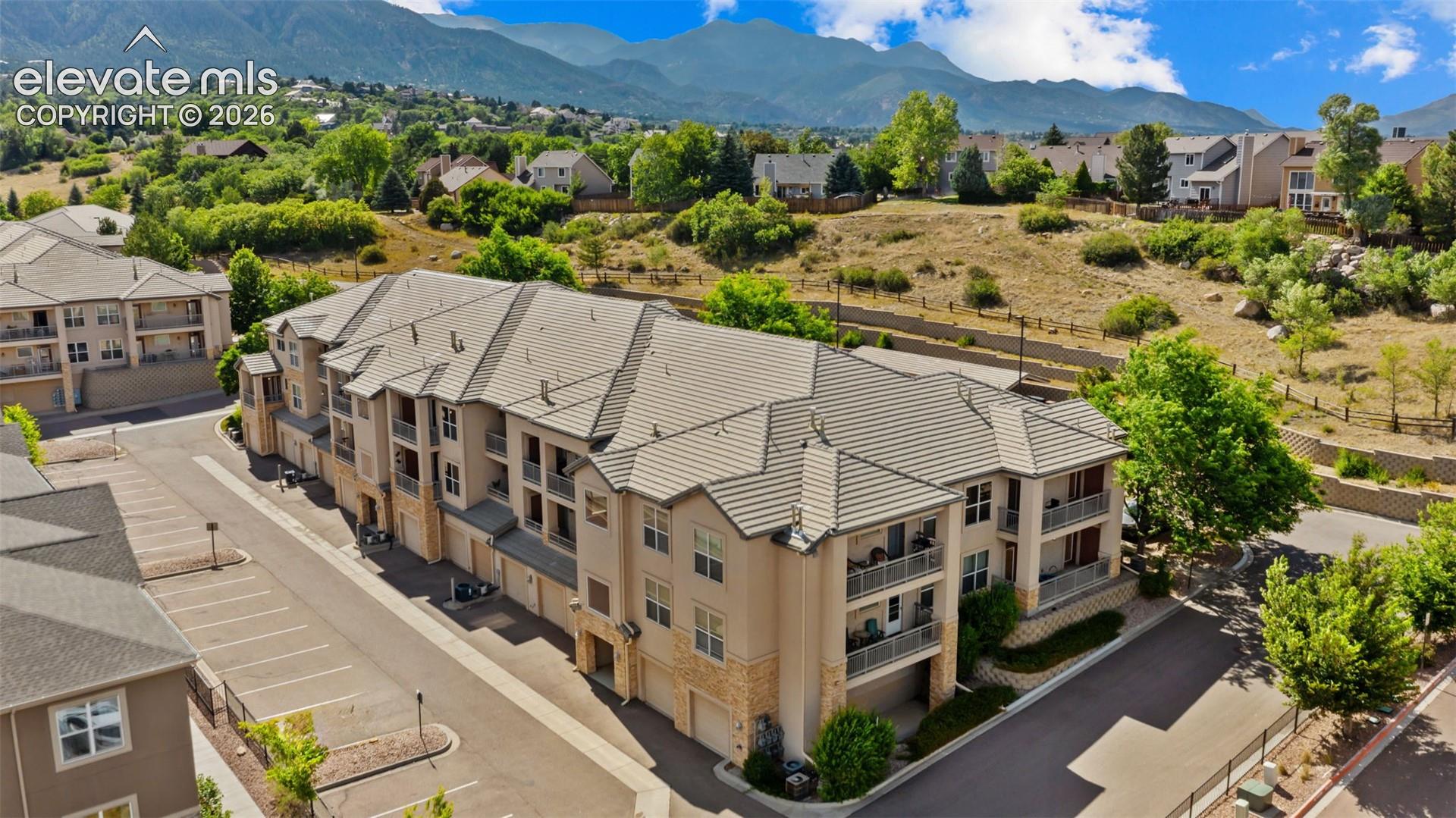 Multi-story residential building featuring a neutral-toned stucco and stone facade, private balconies, covered parking garage entrances, a tiled roof, and a paved parking lot