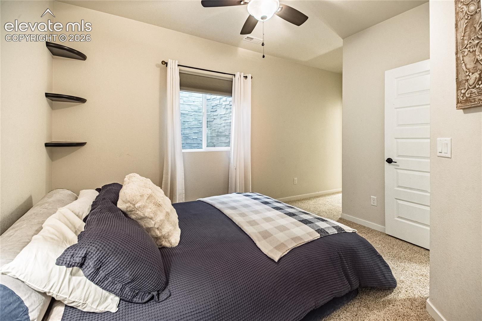 Wall-mounted floating shelving, ceiling fan with integrated lighting, carpeted flooring, and a five-panel interior door