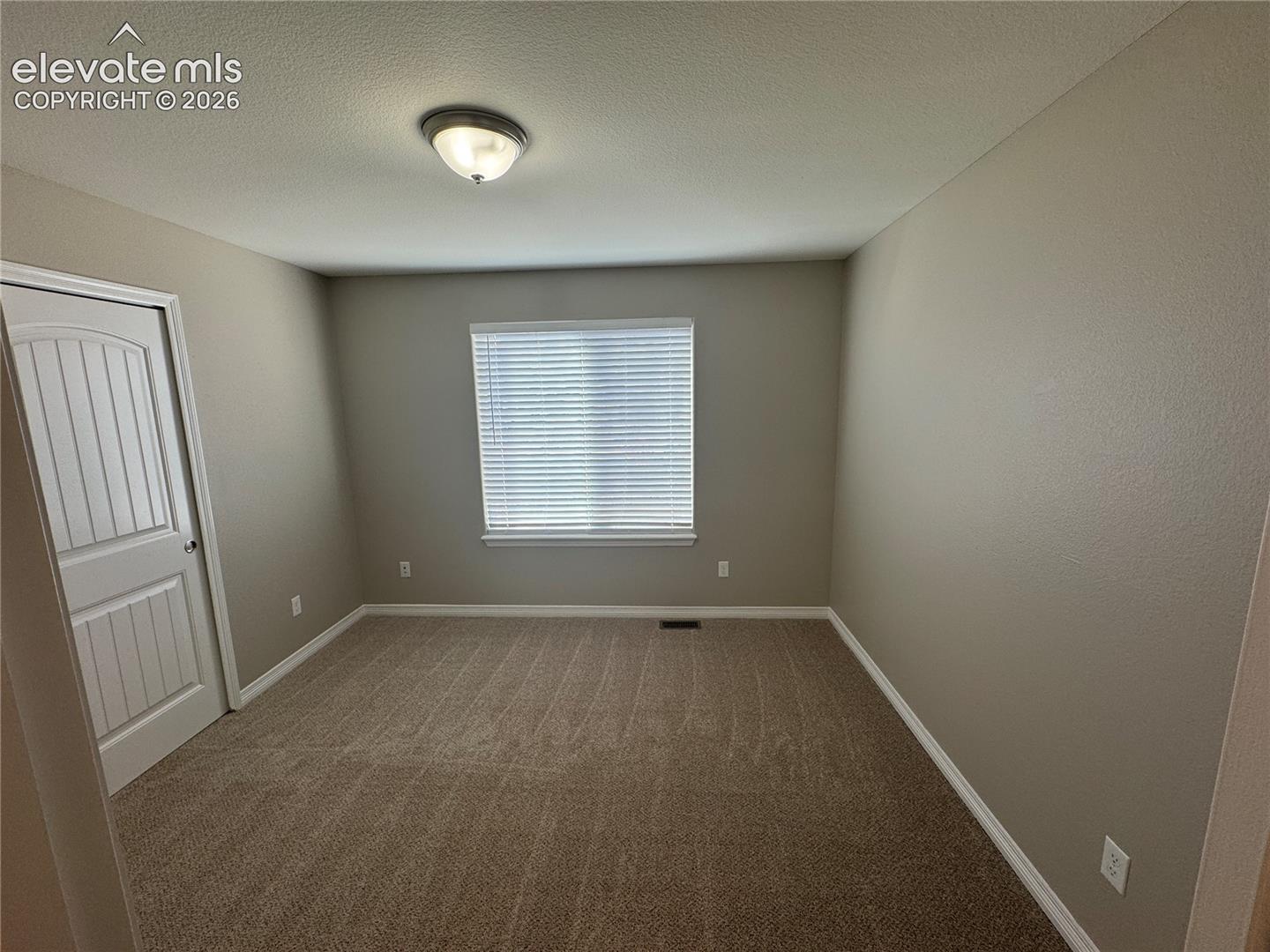 Neutral-toned walls, wall-to-wall carpeting, single-panel interior door with arched detail, flush-mount ceiling light fixture, and window with horizontal blinds