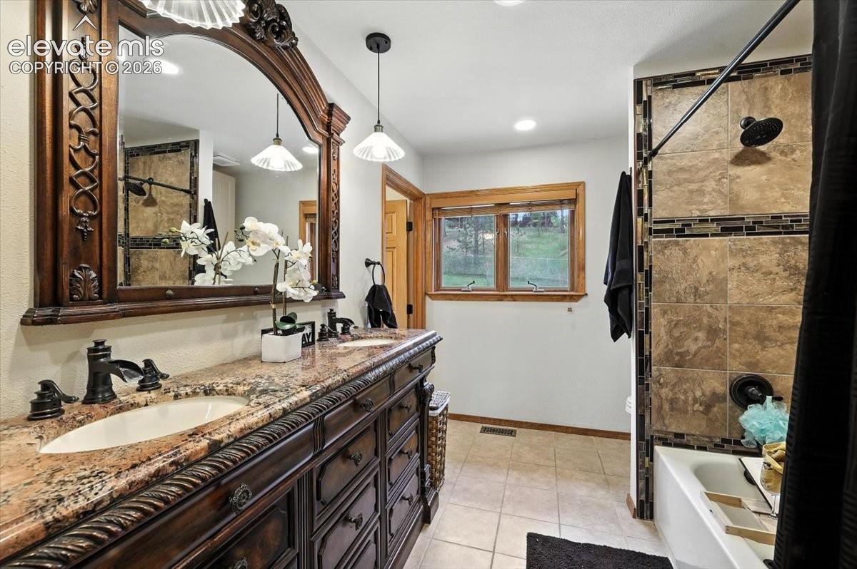 Bathroom featuring a dark wood-finish vanity with dual integrated sinks and stone-patterned countertops, a large ornate-framed wall mirror, pendant lighting, and a shower-tub combination with tile surround and decorative inlay