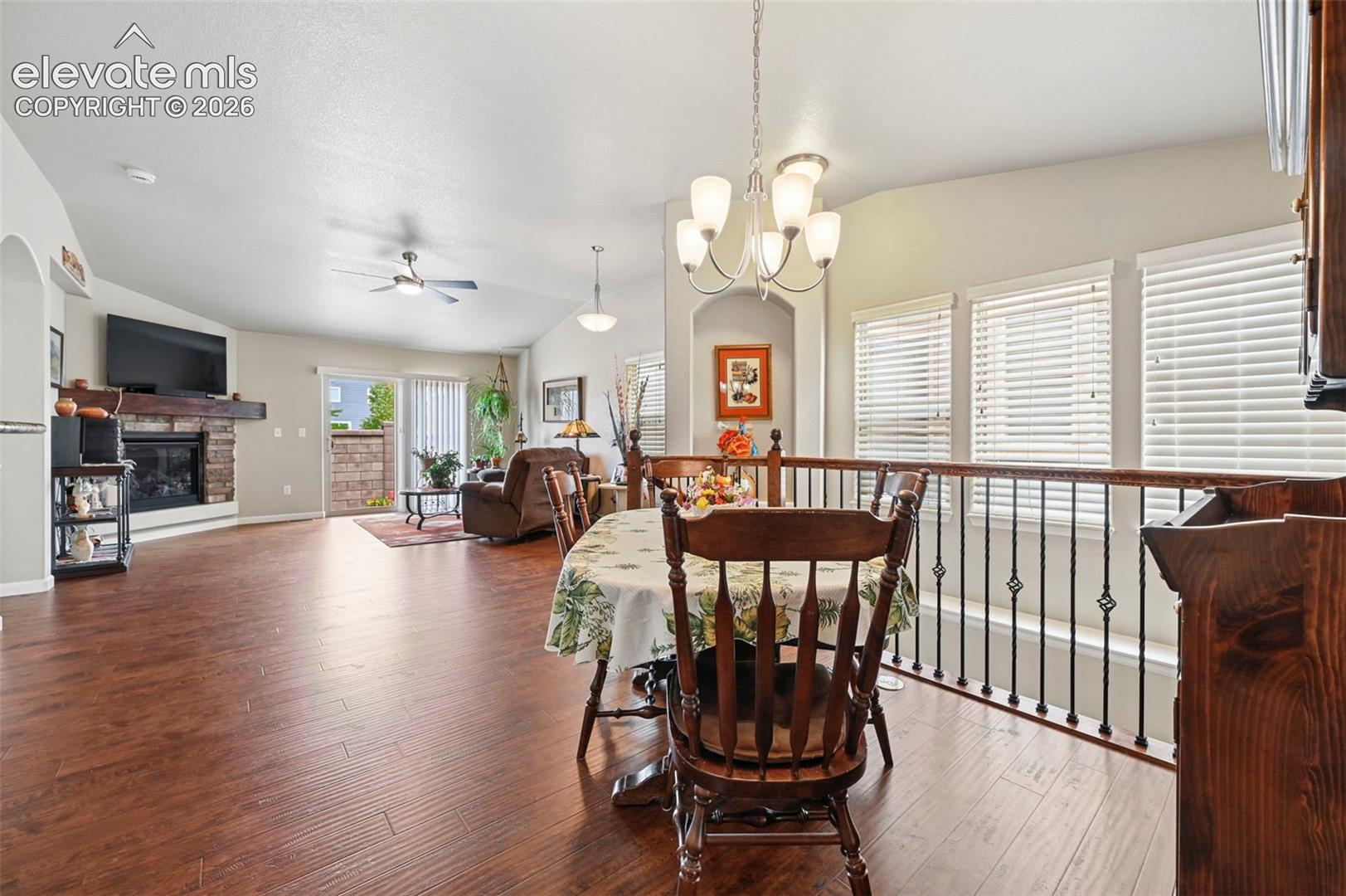 Open-concept living space featuring wood-finish flooring, a stone fireplace with a substantial wood mantel, and vaulted ceilings with a ceiling fan