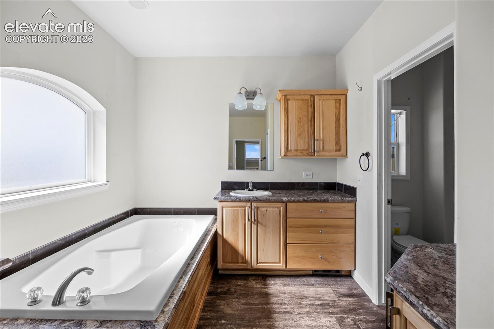 Built-in vanity featuring natural wood cabinetry and stone-finish countertops, arched window, deep soaking tub with deck-mounted faucet, and two-light wall sconce