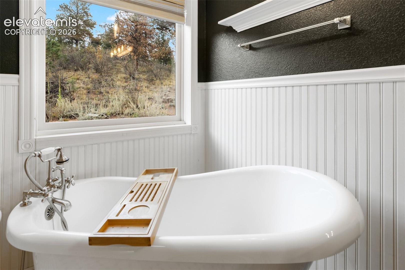 Freestanding soaking tub featuring a silver-tone faucet with a handheld shower attachment, beadboard wainscoting, and a window with exterior scenery