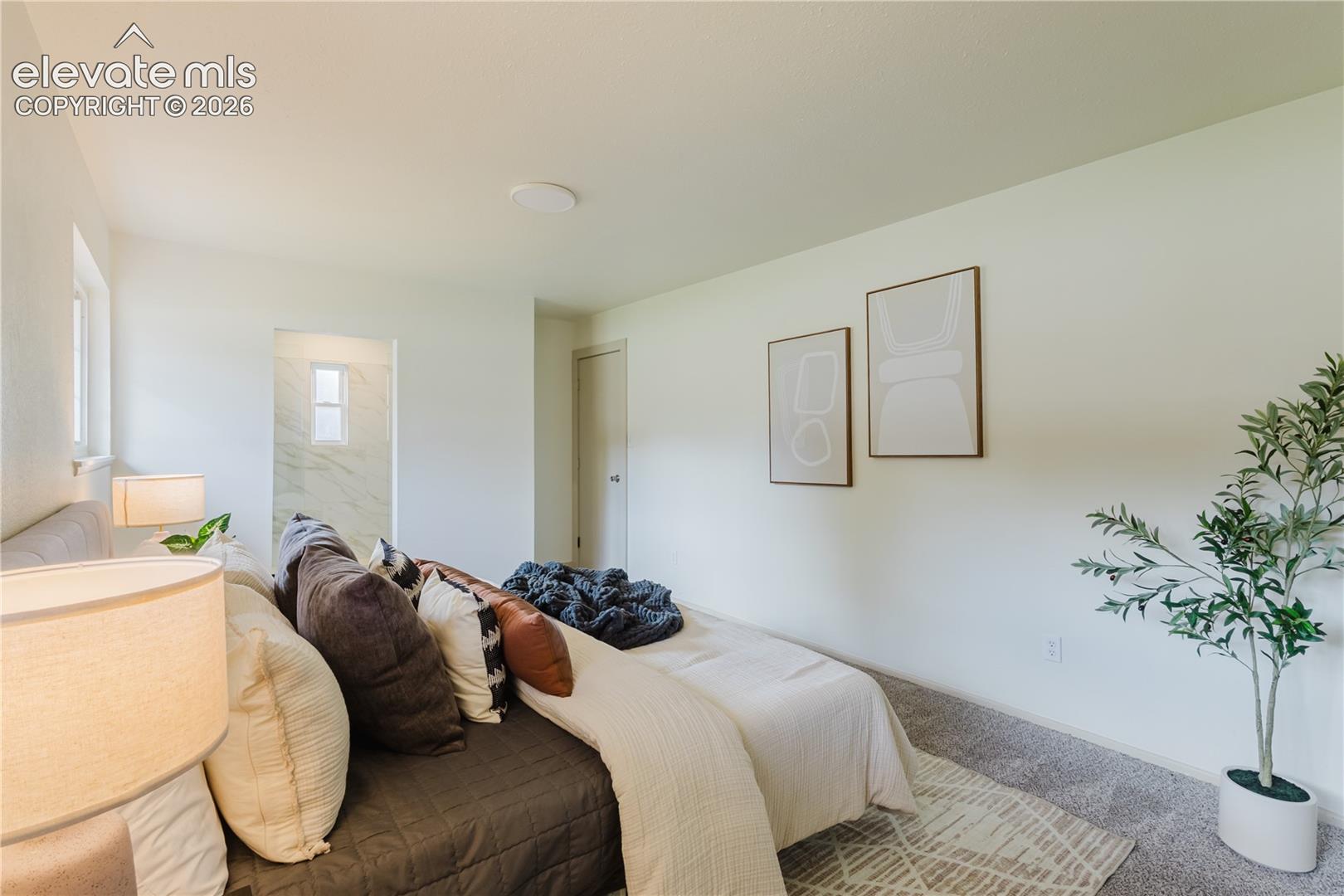 Carpeted bedroom with white walls, recessed ceiling lighting, and a doorway opening to a bathroom featuring a window and marbled wall tiling