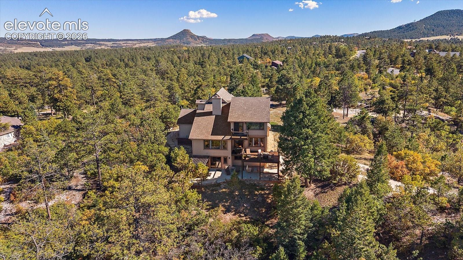 Multi-level structure featuring a multi-pitched shingled roof, exterior chimney, raised wooden deck, and mature evergreen surroundings set against a mountain backdrop