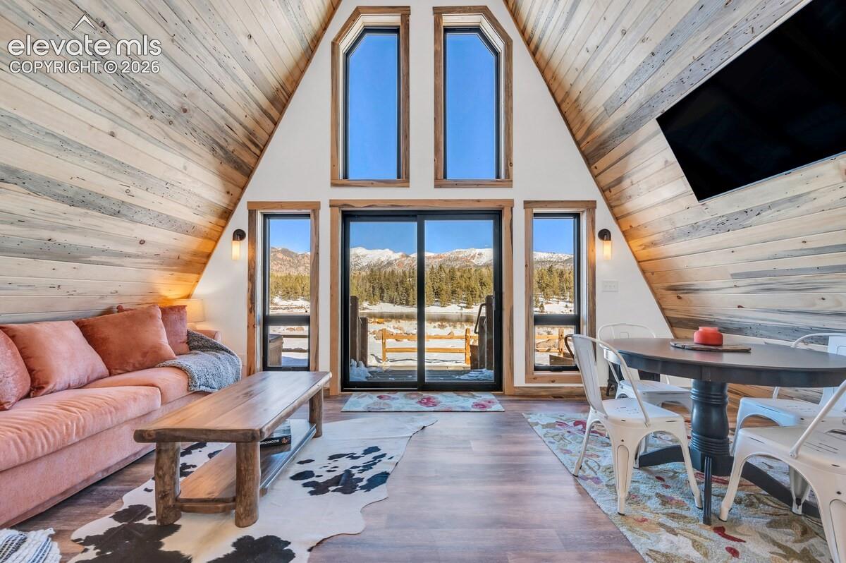 A-frame living space featuring vaulted wood-paneled walls, floor-to-ceiling windows, sliding glass door, wood-finish flooring, and wall-mounted light fixtures