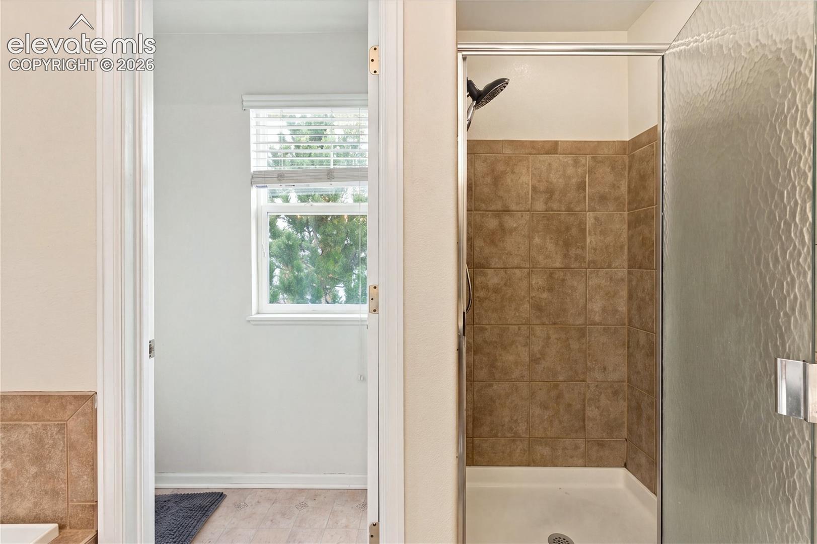 Walk-in shower with textured glass enclosure, floor-to-ceiling neutral tile surround, and window with adjustable blinds
